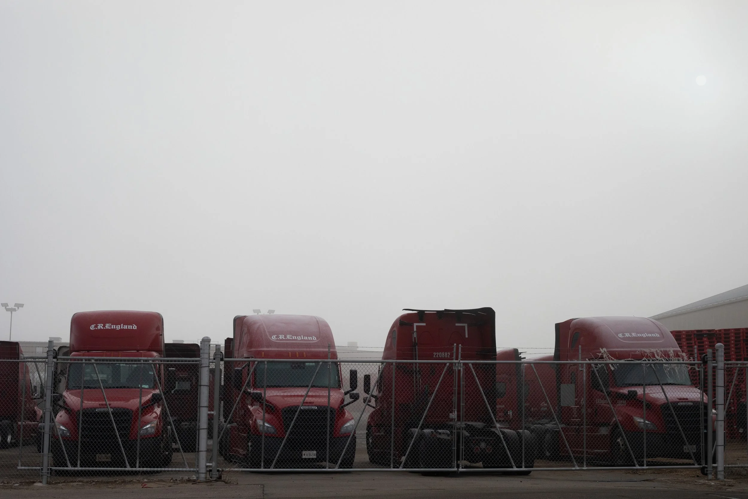 C.R. England trucks behind chain-link, winter fog, Denver, CO