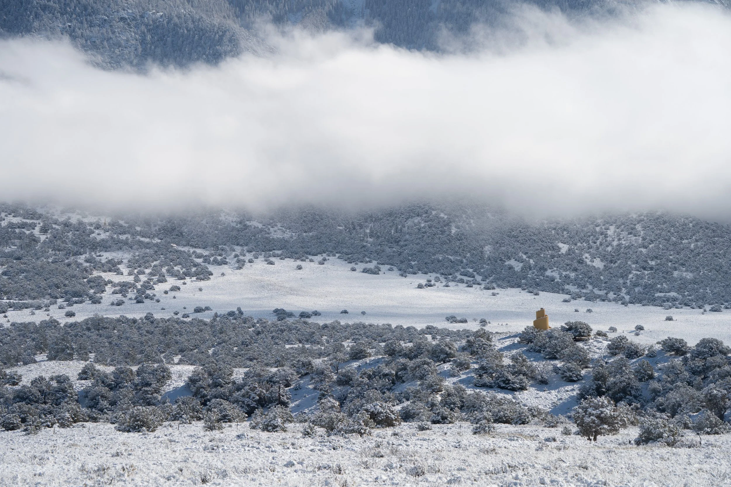 Snow-covered hillside and scrub trees below a low cloud bank, ziggurat in middle distance, Crestone, CO