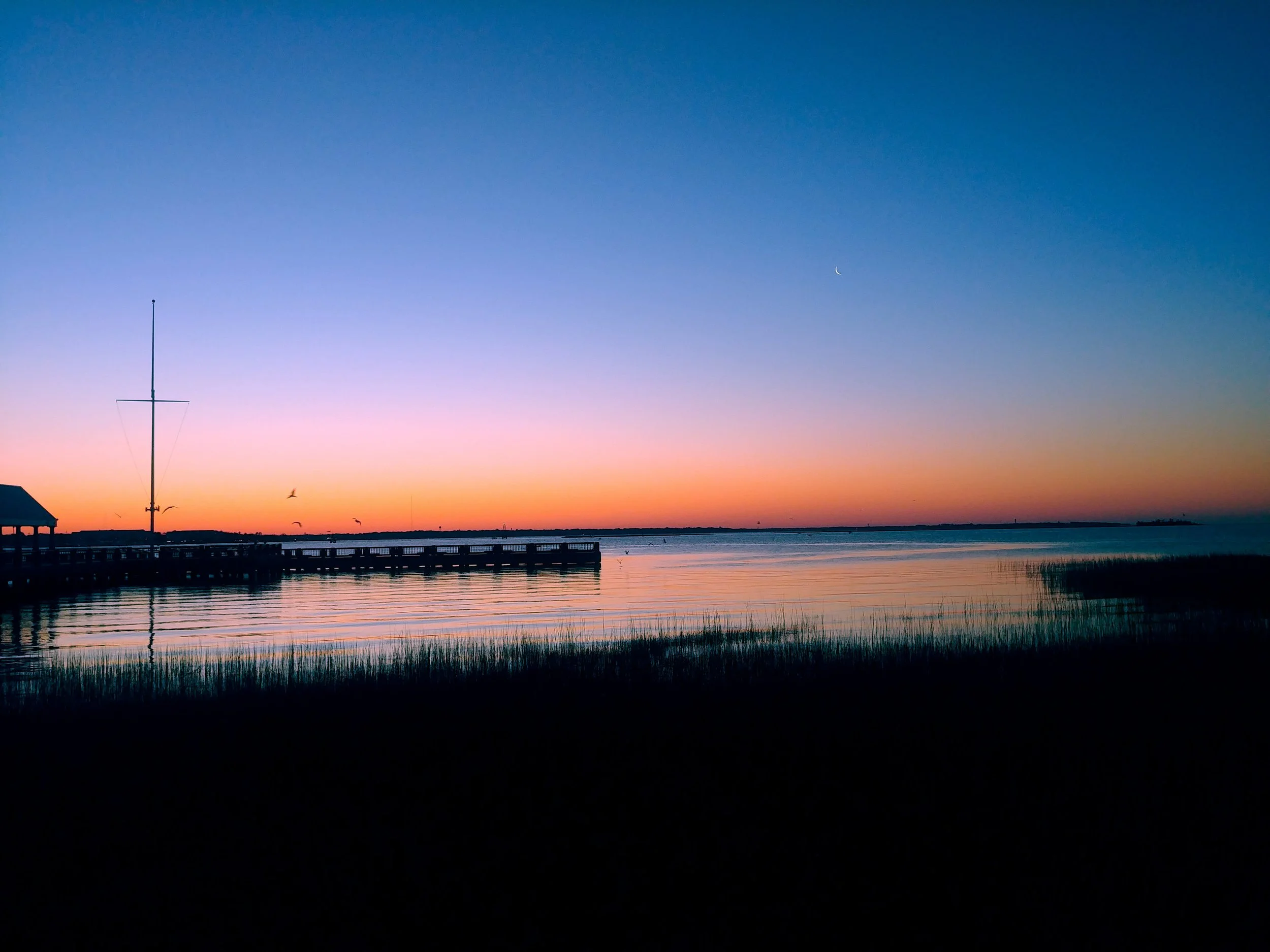 Coastal sunset with dock, mast, marsh grass, and crescent moon reflected on calm water, Charleston, SC