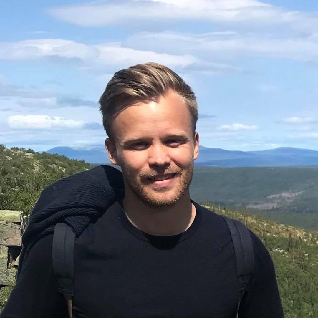 En man med kort, lätt lockigt hår och skägg, bär svart tröja och ryggsäck, står utomhus med landskap av kullar och blå himmel i bakgrunden.