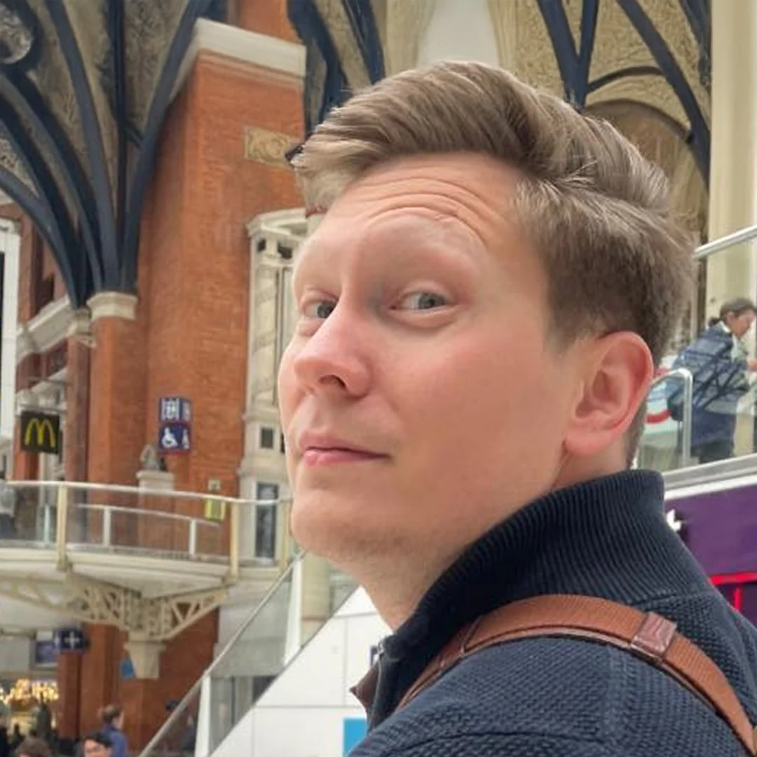 En man med ljusbrunt, tvåfärgat hår i en träningdräkt, som ser bakåt mot kameran, i en inomhusmiljö med arkitektoniska element av gult tegel och blå detaljer.