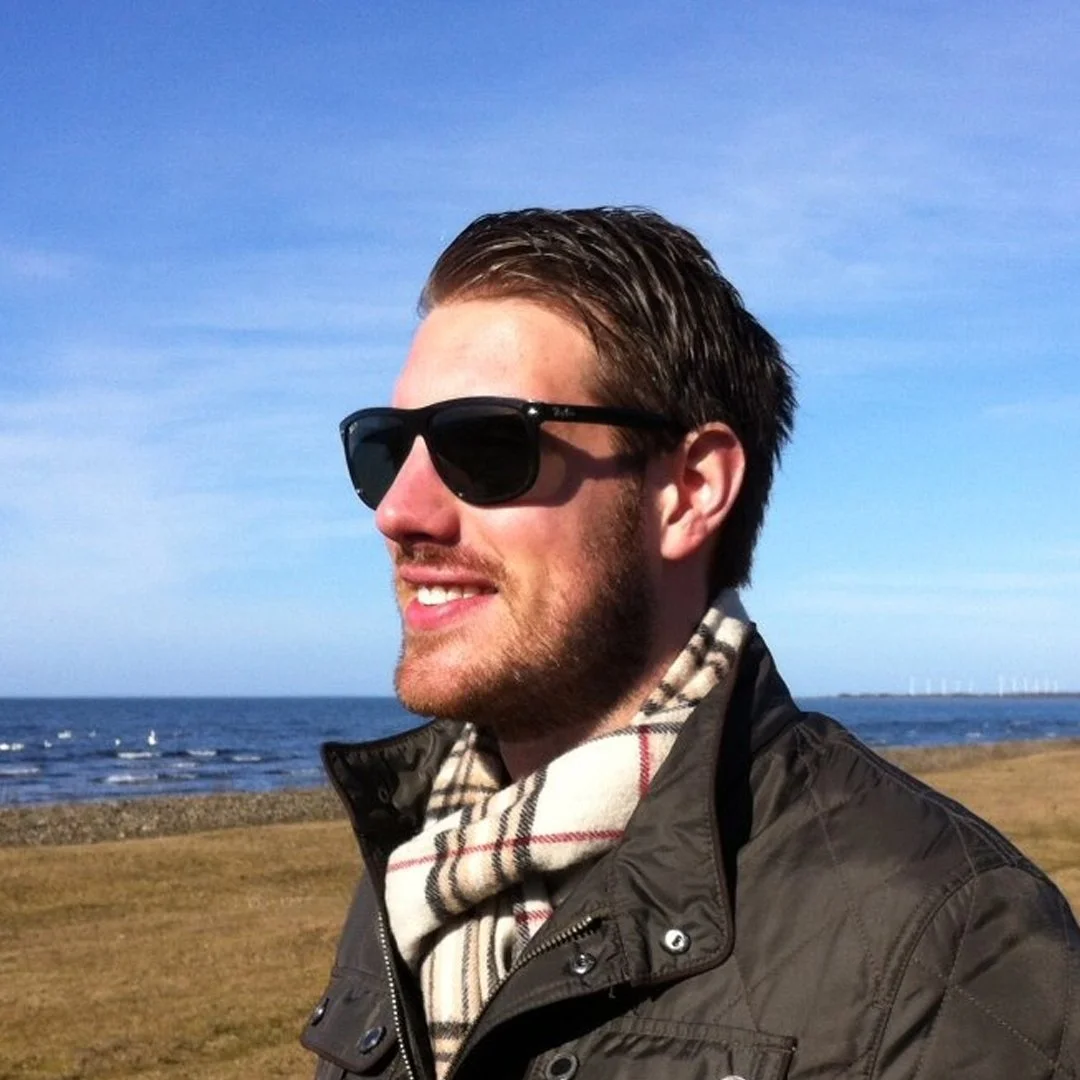 Man i solglasögon, jacka och halsduk står vid havet med blå himmel och vindkraftsverk i bakgrunden.