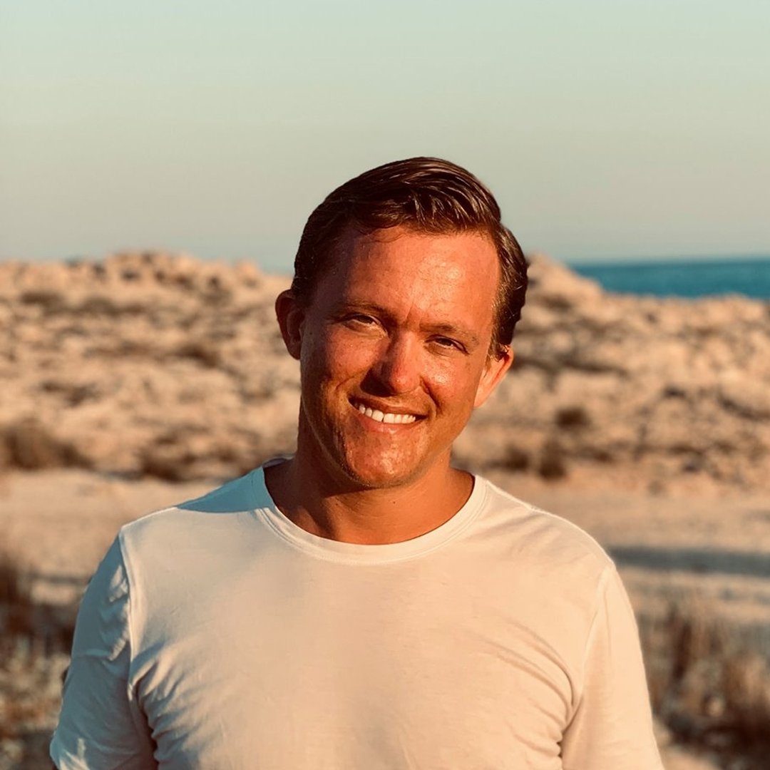 En man med brunt hår och vit T-shirt står på en strand med stenar och hav i bakgrunden genom solsken.