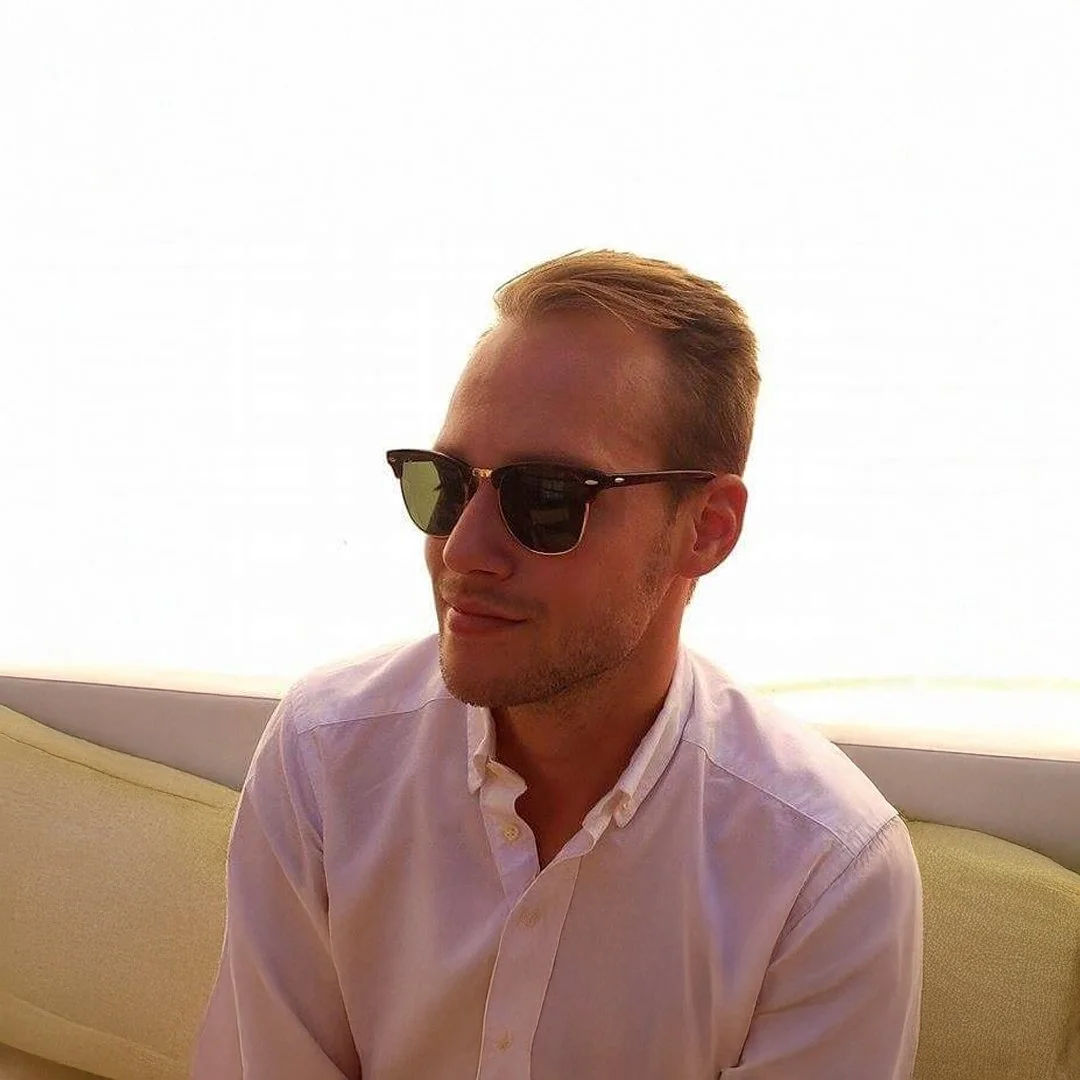 Ung man med solglasögon och vit skjorta, sittande mot en ljus bakgrund.