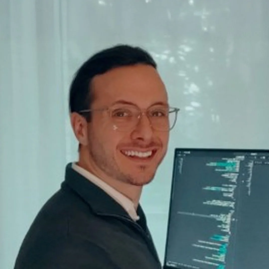 En man med glasögon sitter framför en dator med kod på skärmen och ler.