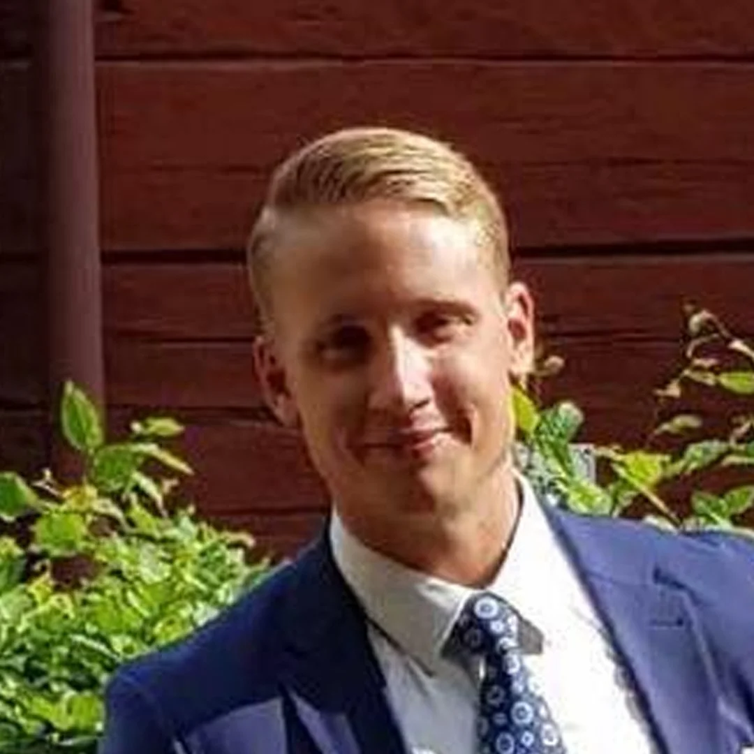 En man i kostym står framför ett trävägg och gröna växter.