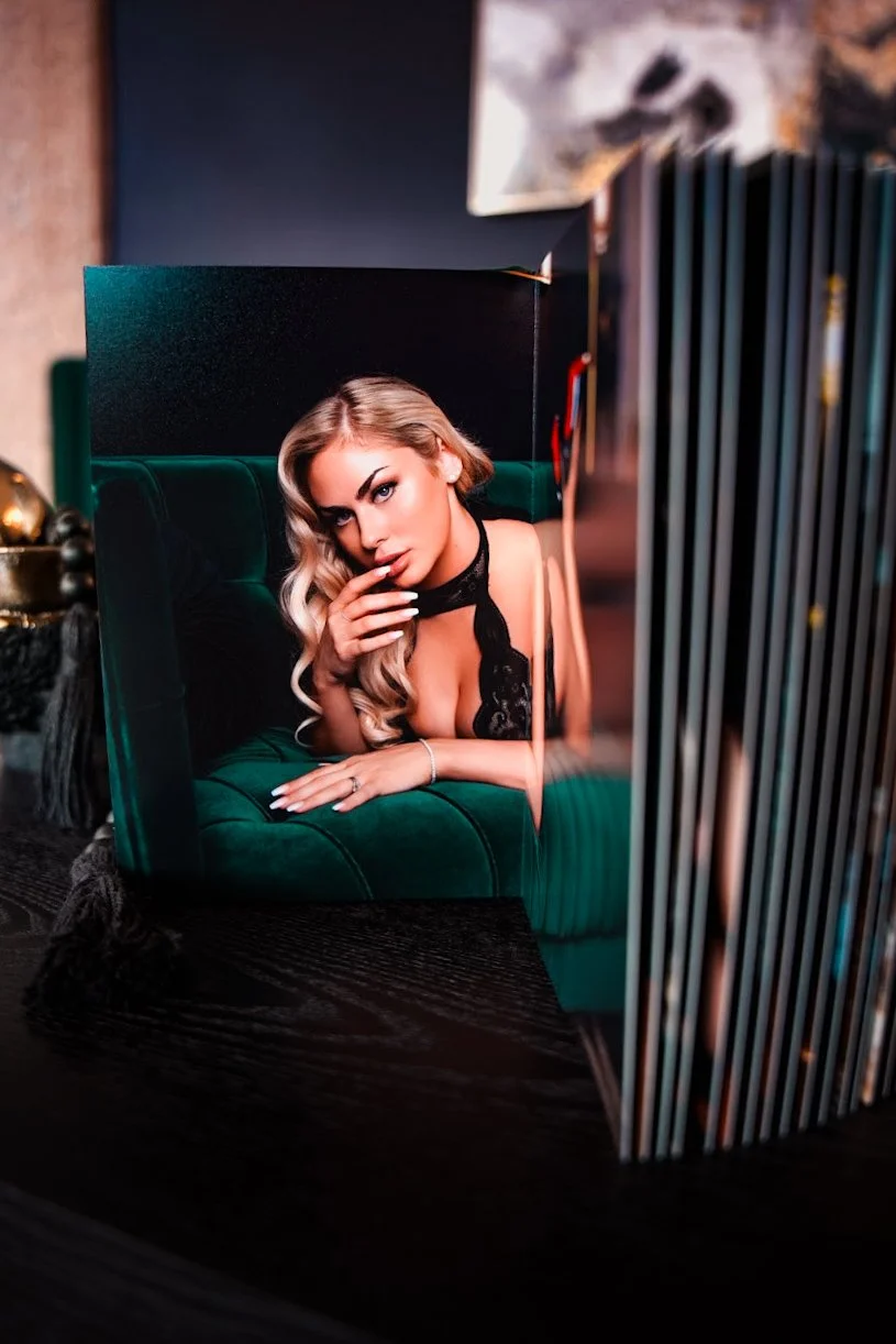A photograph of a woman with blonde hair styled in loose curls, wearing a black lace top, posed on a green velvet couch with her hand near her face. The image is printed on a piece of paper that is standing upright, with other similar prints or objec
