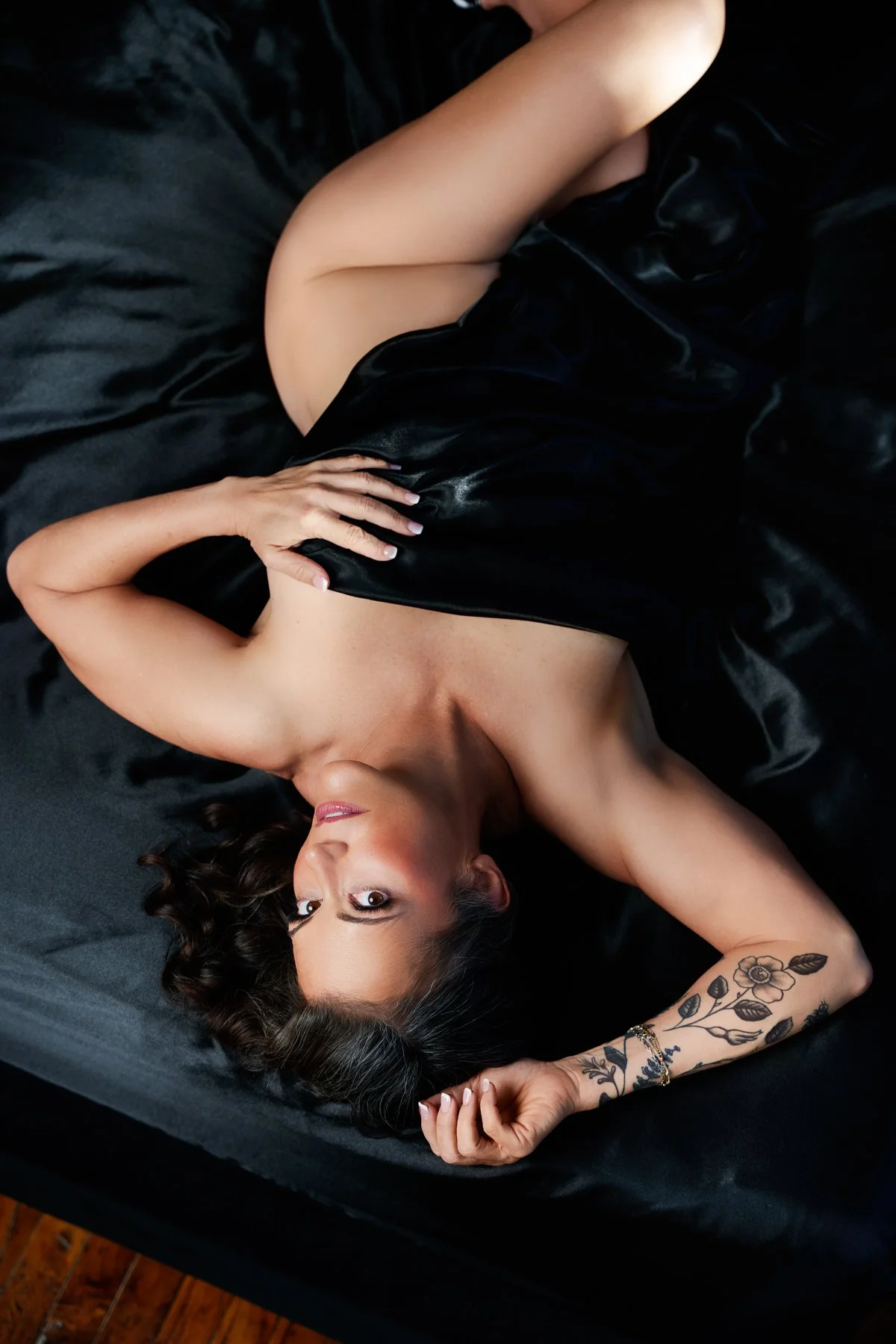 A woman lying on a black bed with black sheets, wearing a black satin dress, with her head turned to the side and her eyes looking up. She has a flower tattoo on her left forearm and is holding a part of her dress with her left hand, while her right 