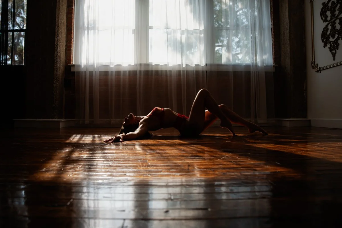 A person in lingerie lying on their back on a wooden floor with sunlight streaming through sheer curtains on a large window.