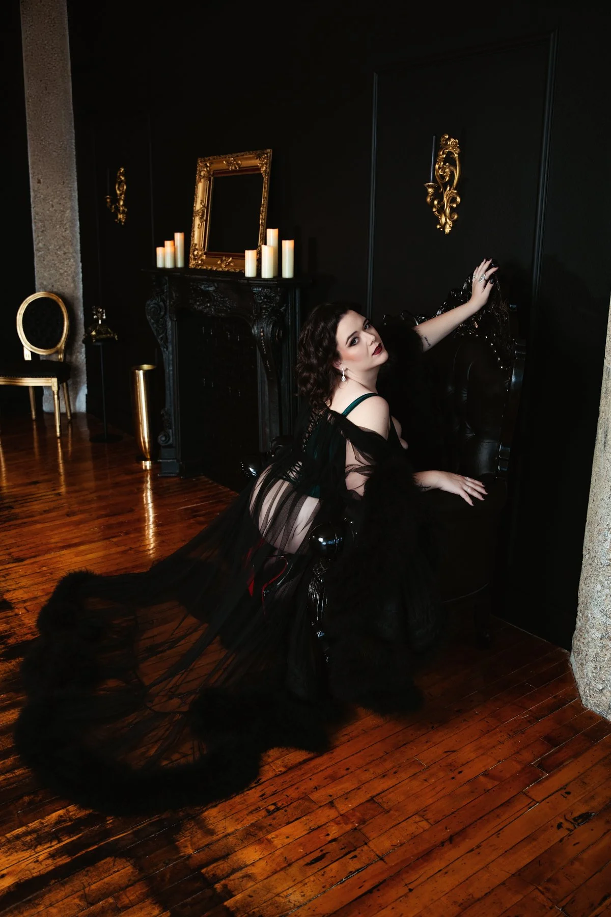 A woman dressed in black, sitting on a black chair with her legs extended, leaning against a black wall with ornate gold decorations and candles on a black mantle behind her. She has dark hair, makeup, and wears jewelry, with a sheer black gown trail