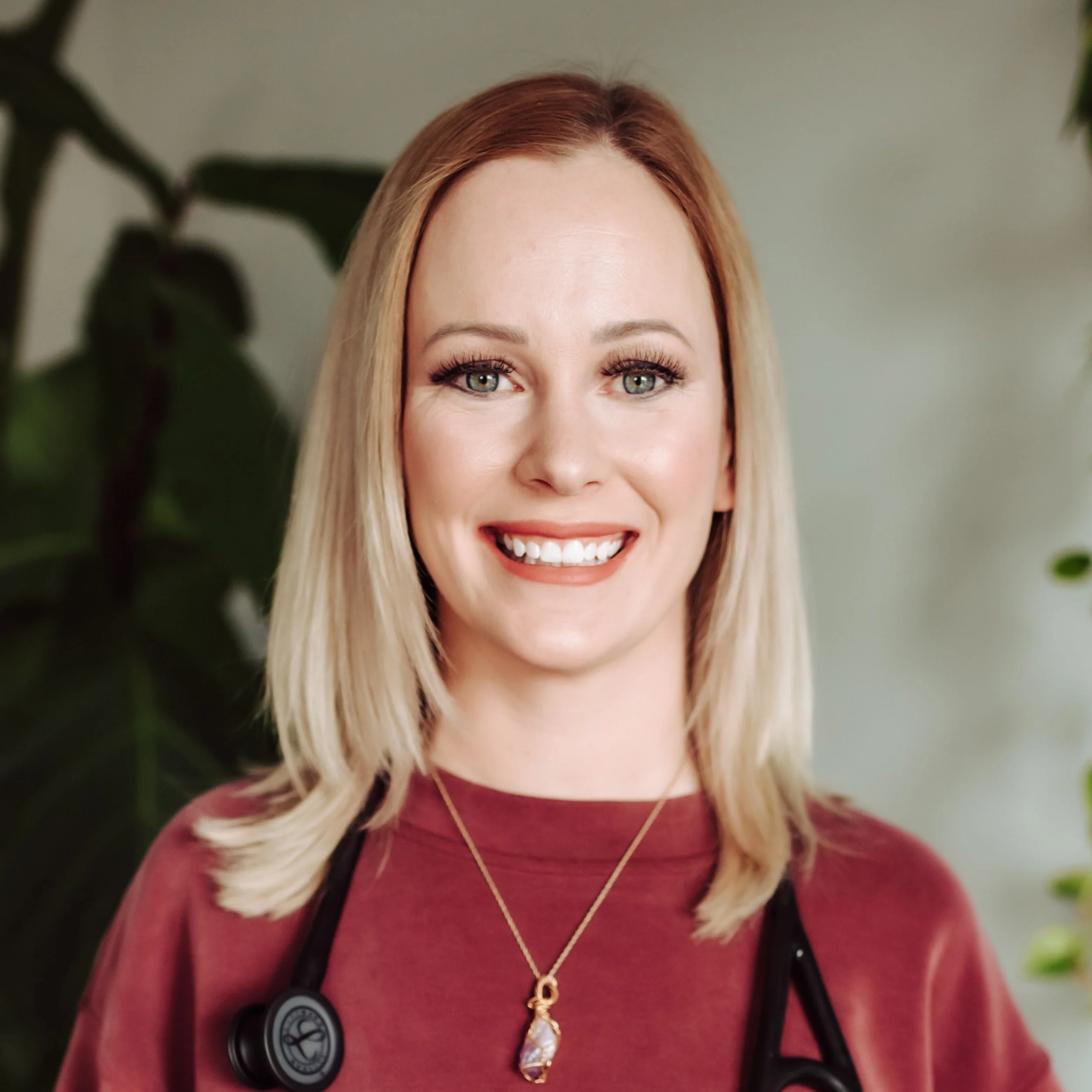 A smiling woman with blonde hair wearing a maroon top and a gold necklace, with a stethoscope around her neck, standing in front of green plants.