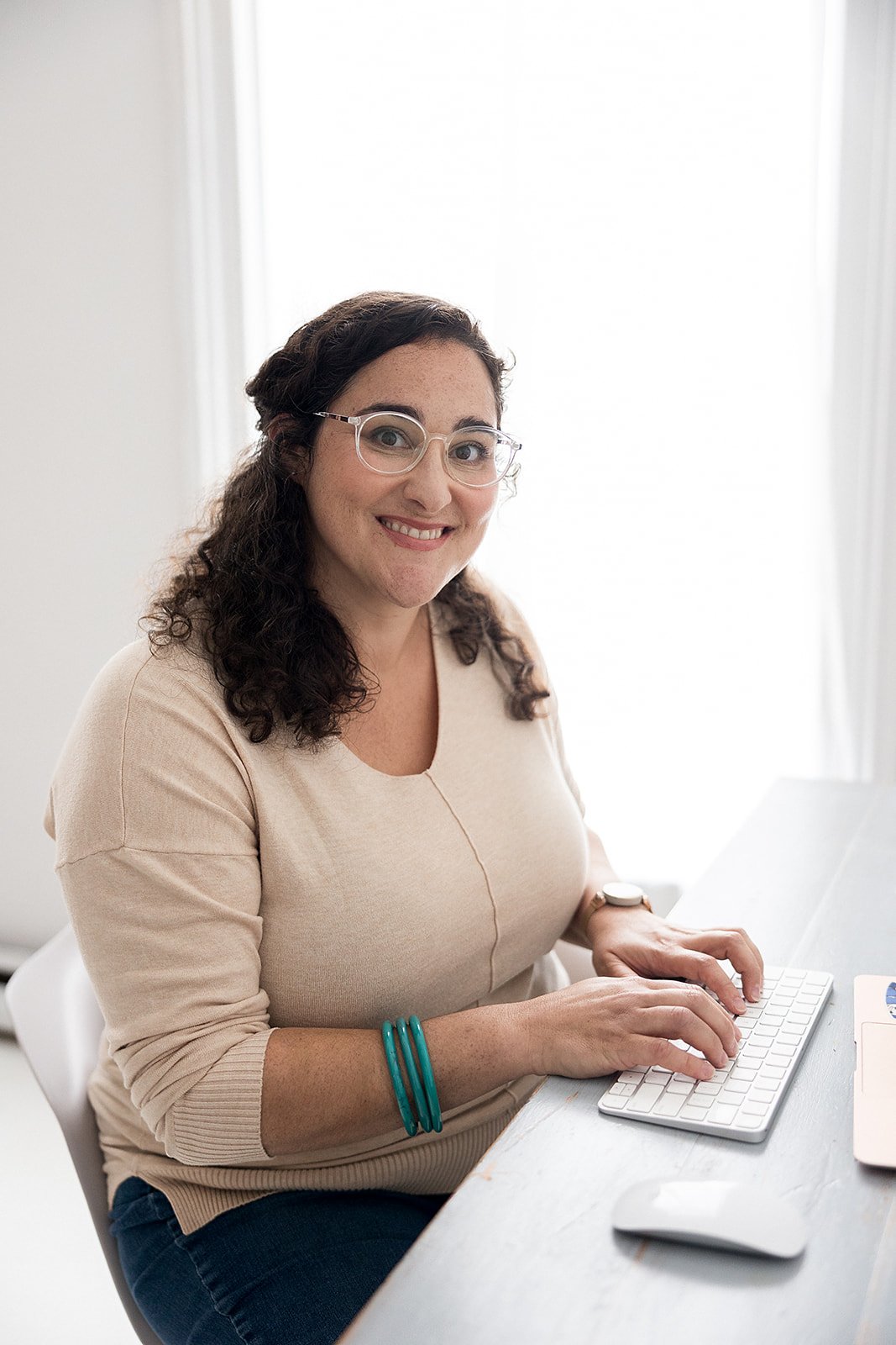 A woman with dark, curly hair, wearing glasses, a beige top, and blue bracelets, smiling while sitting at a desk with a keyboard and mouse in front of her, in a well-lit room with sheer white curtains.