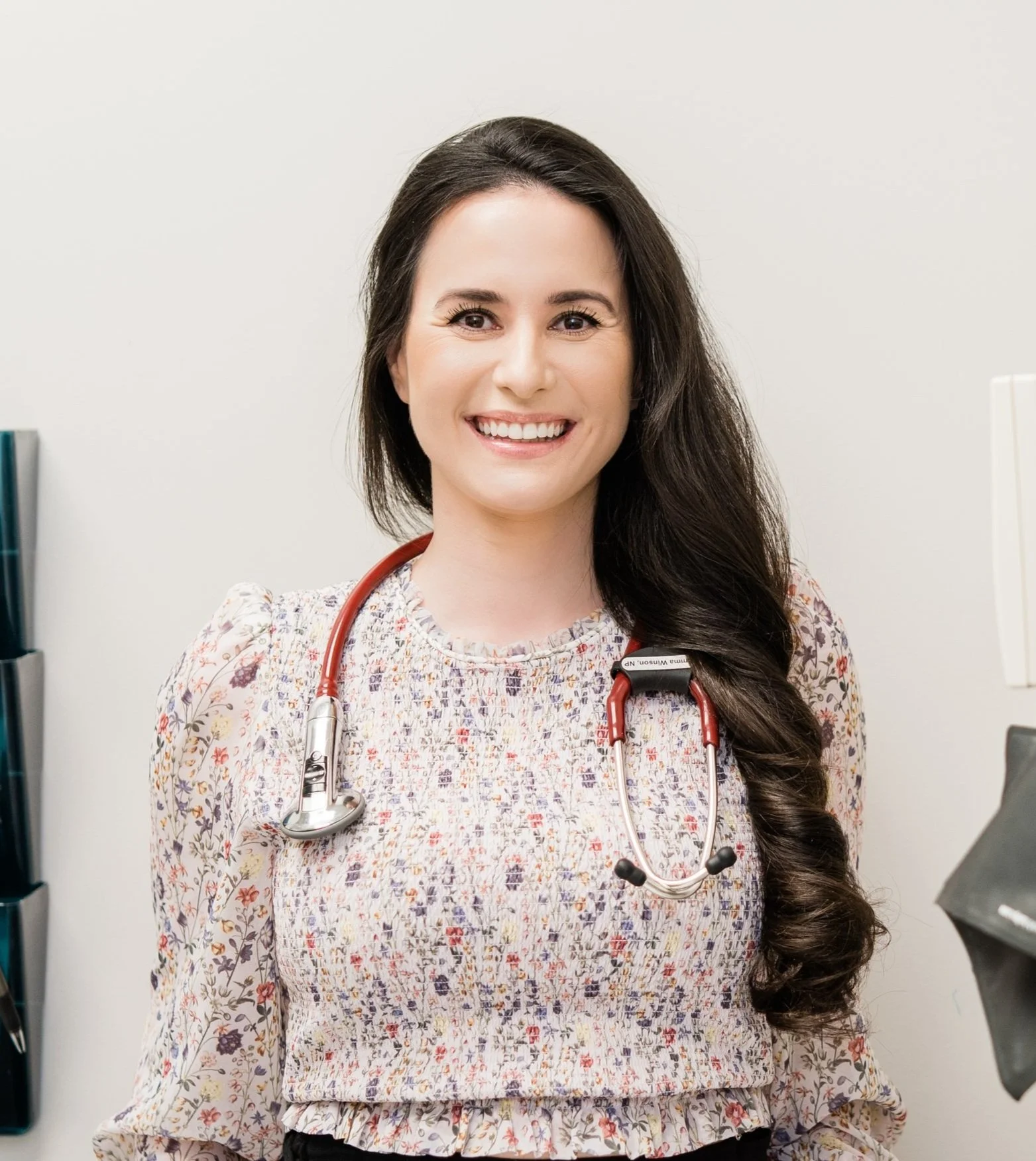 Smiling female doctor with long dark hair, wearing a floral blouse and red stethoscope, standing in a medical office.