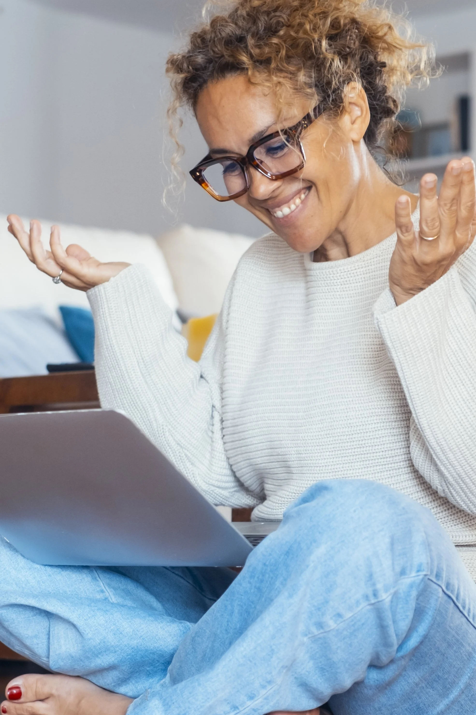 A smiling woman with curly hair and glasses sitting on the floor, looking at her laptop with a joyful expression.