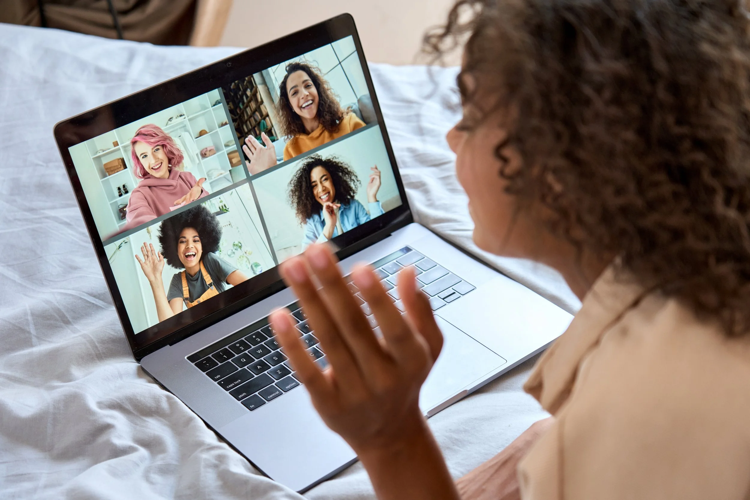 Woman with curly hair participating in a video call with four women, each shown in a separate window on her laptop screen, all smiling and waving.