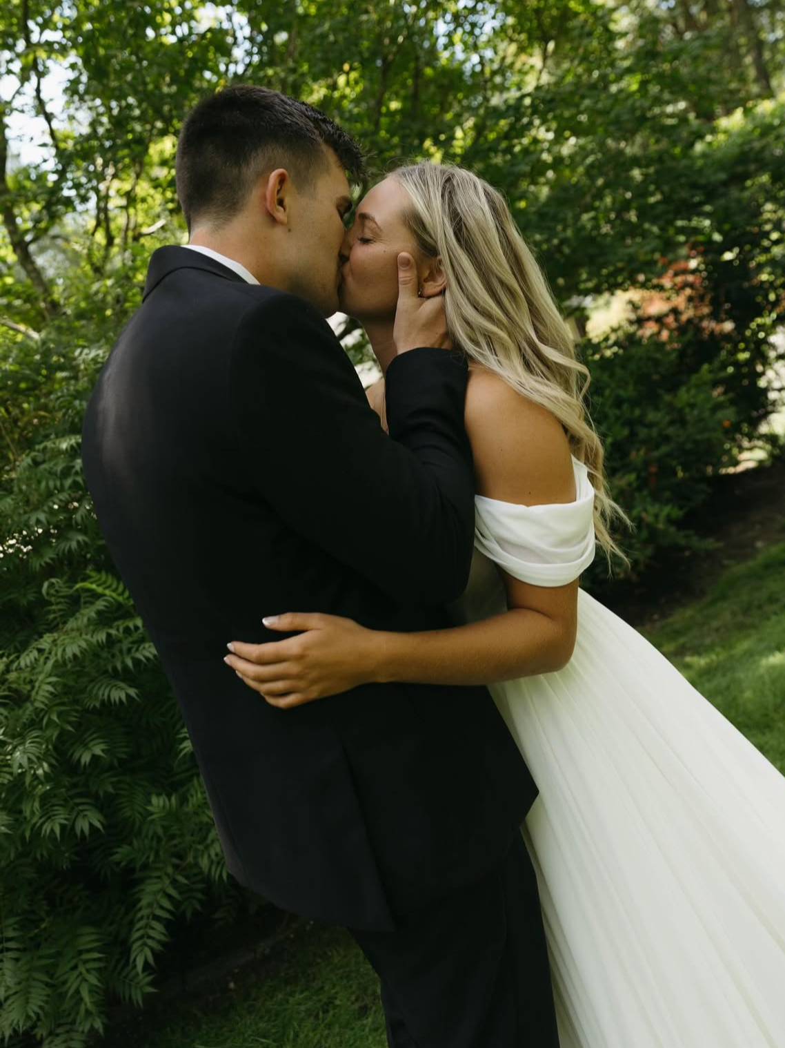 A couple in wedding attire shares a kiss outdoors surrounded by green trees and foliage.