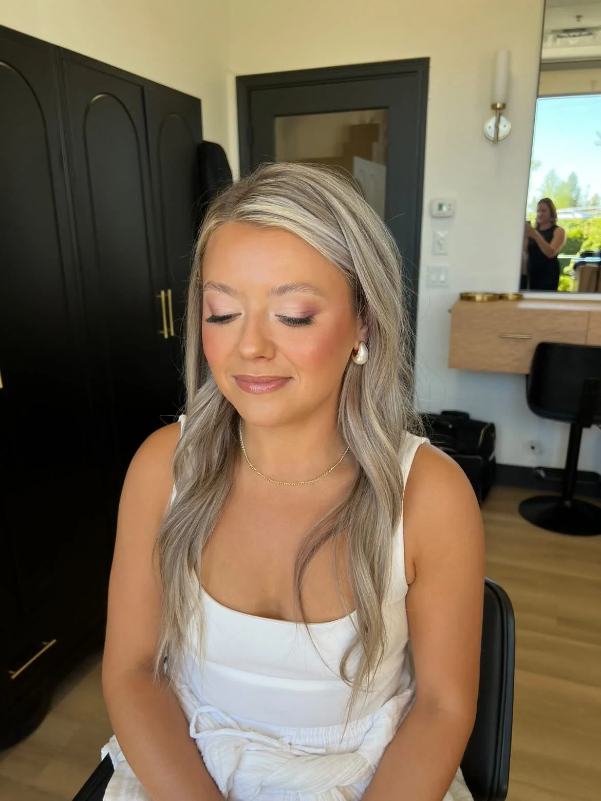 A woman with long, wavy blonde hair and pearl earrings sitting with eyes closed in a room with black cabinets, a mirror, and a person taking a photo in the background.