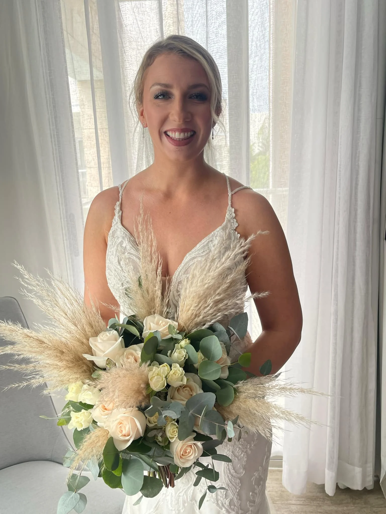 A smiling bride in a white lace wedding dress holding a bouquet of white roses, greenery, and pampas grass.