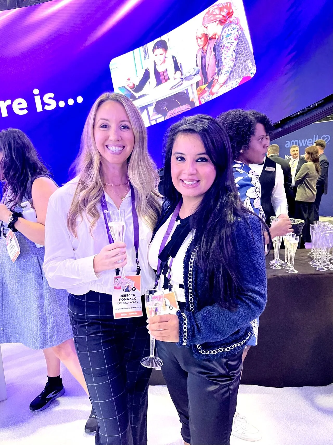 Two women at a professional event holding glasses of champagne, with a large blue screen in the background displaying the words 'There is...' and a photo of people working together.