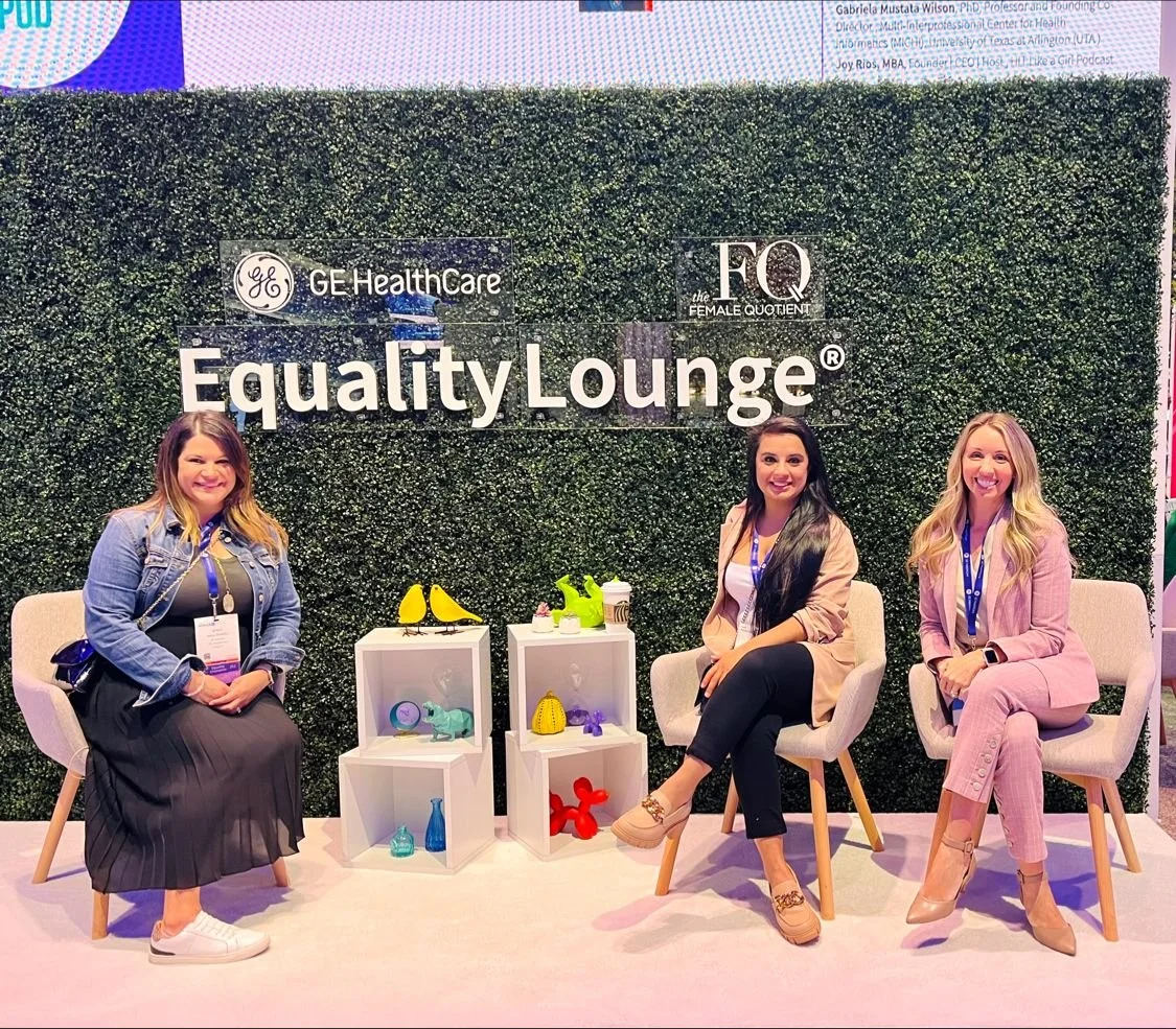 Three women sitting on chairs in front of a green wall with the signage 'Equality Lounge' and logos for GE Healthcare and The Female Quotient at a conference or event. Small display shelves with colorful decorative objects are between them.
