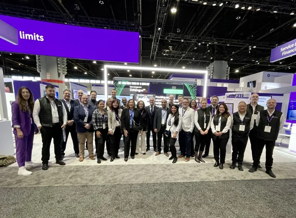 Group of people standing in a conference or expo hall, with booths and digital displays in the background, and a purple sign above that reads 'limits'.