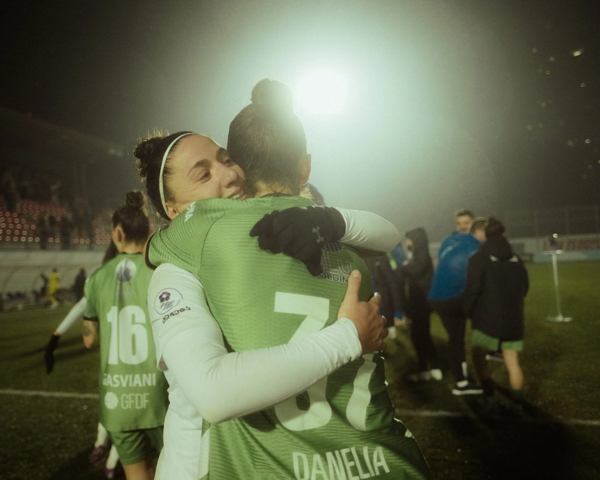 Two football players, one with jersey number 37 and the name DANELIA, hug each other on the field after a match, smiling and celebrating. Other players and people are seen in the background under stadium lights.
