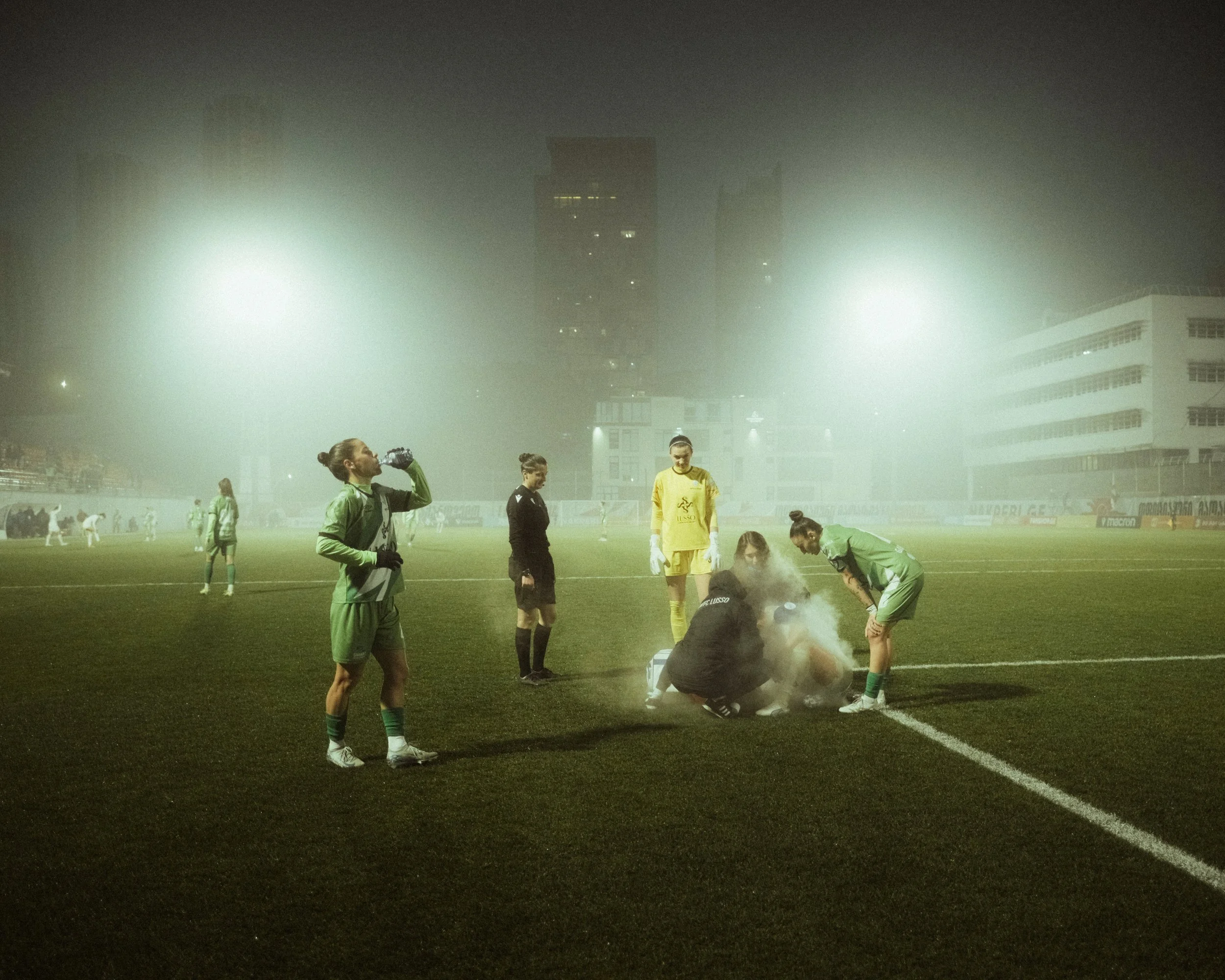 Football players on a foggy field during a game, with one player in black and yellow standing and others attending to a player who appears injured on the ground, surrounded by medical staff, under bright stadium lights.