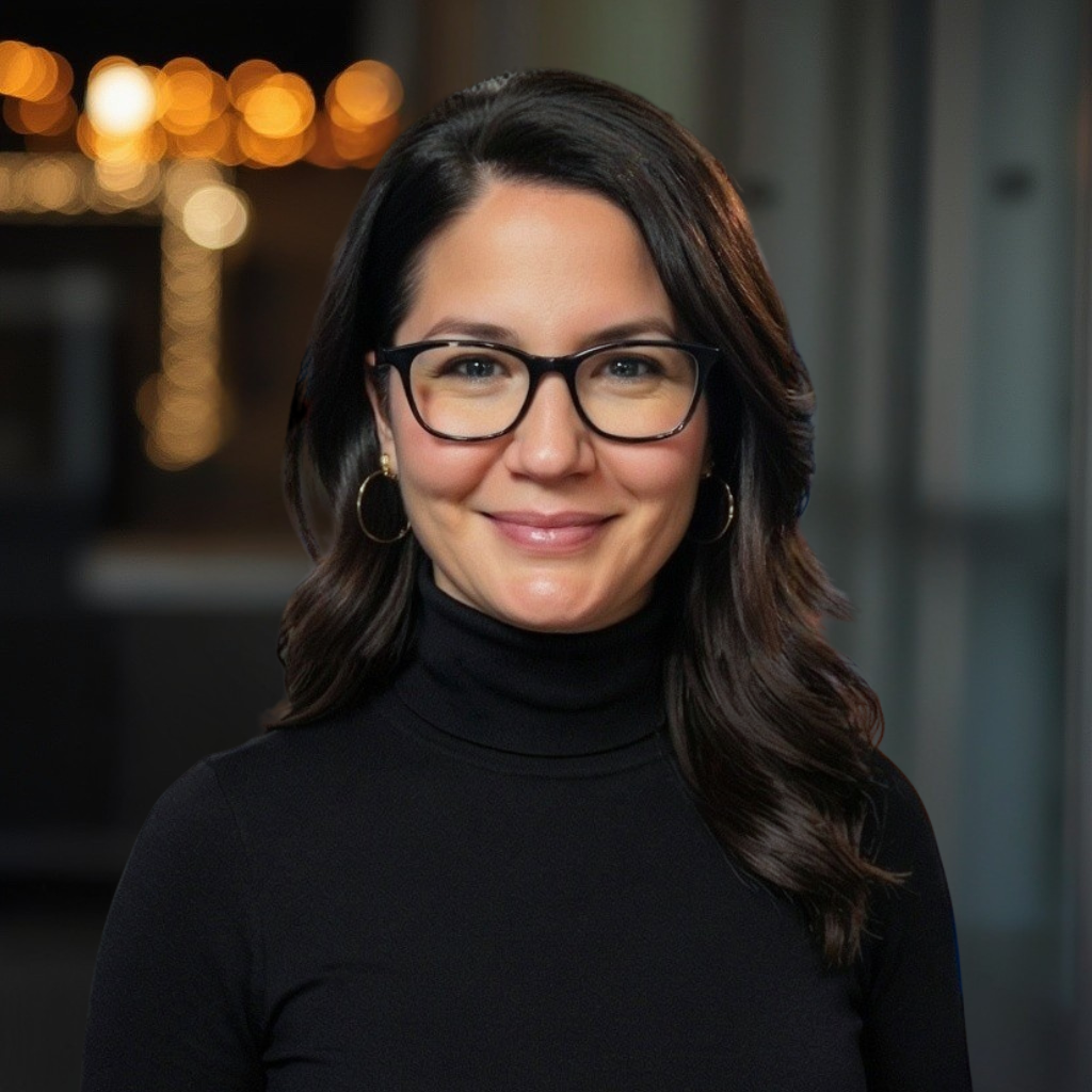 A woman with glasses smiling and dark hair with black clothing and warm backlightin