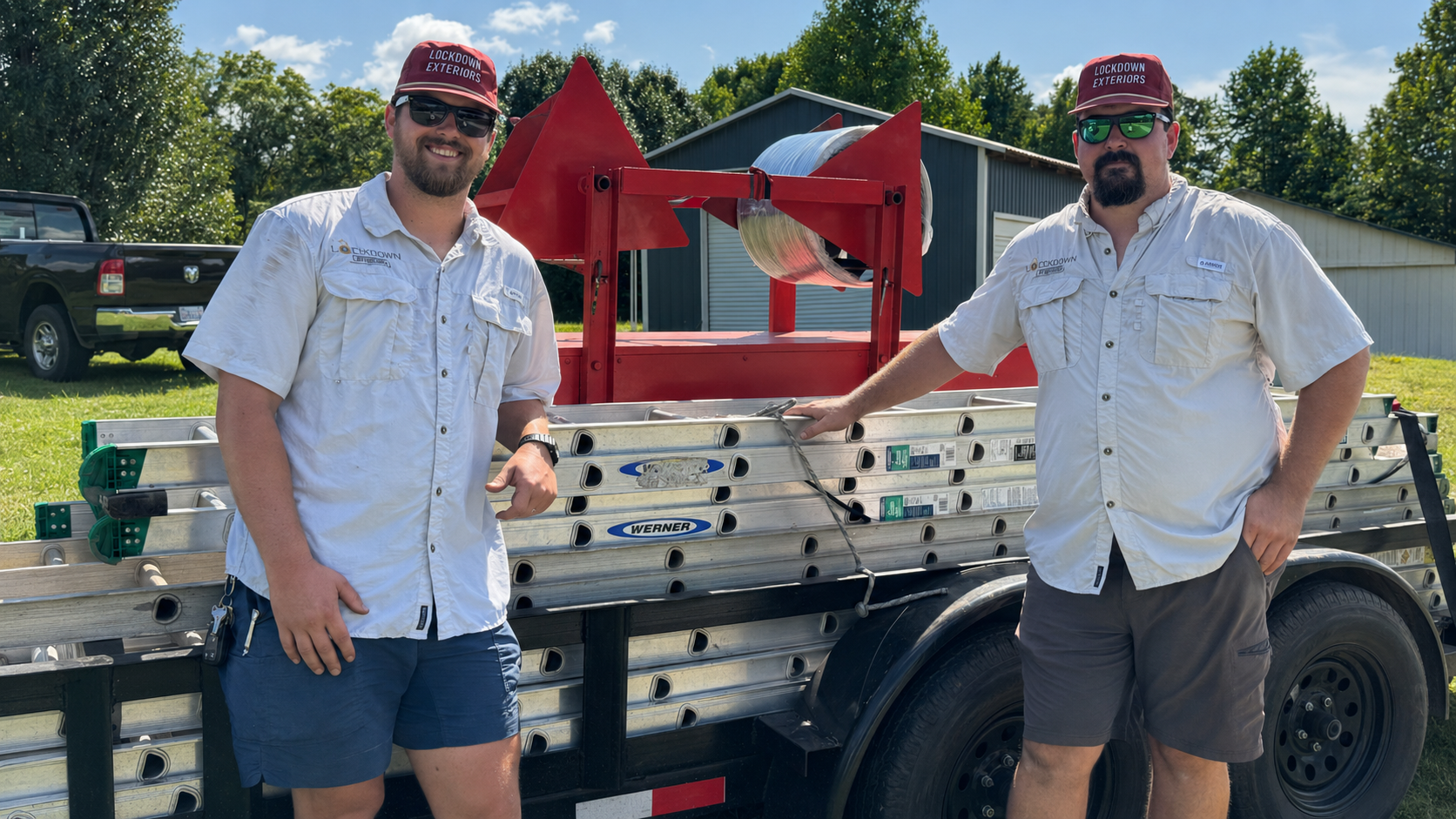 Two men standing by a trailer filled with ladders. One is smiling and wearing sunglasses, the other has a beard and is wearing dark sunglasses. Both are wearing white shirts and red caps that say "Lockdown Exteriors." They are outdoors, with a barn and trees in the background.