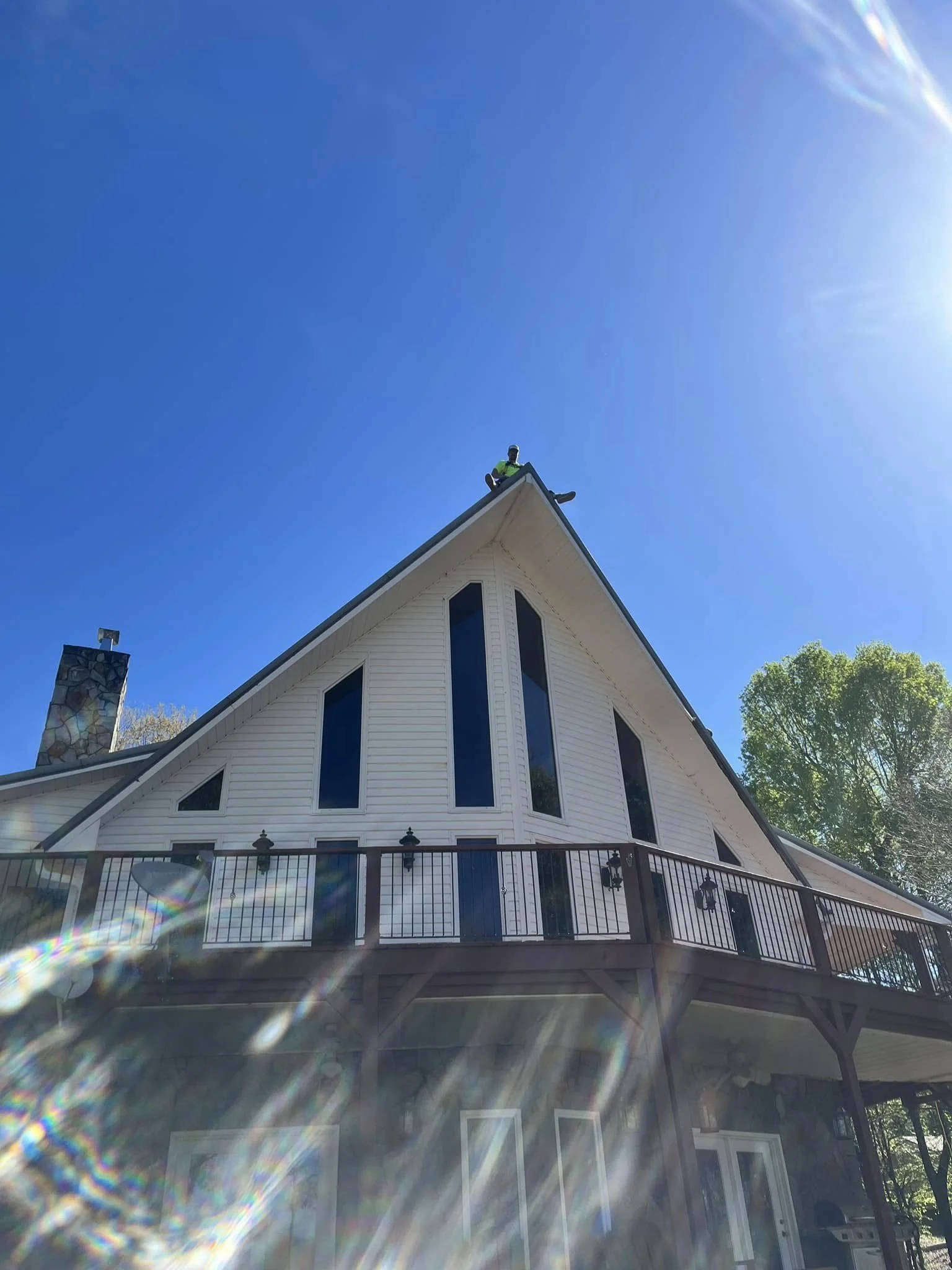 A house with a steeply pitched roof, multiple narrow windows, a chimney, and a balcony, set against a bright blue sky with sunlight flare effects.