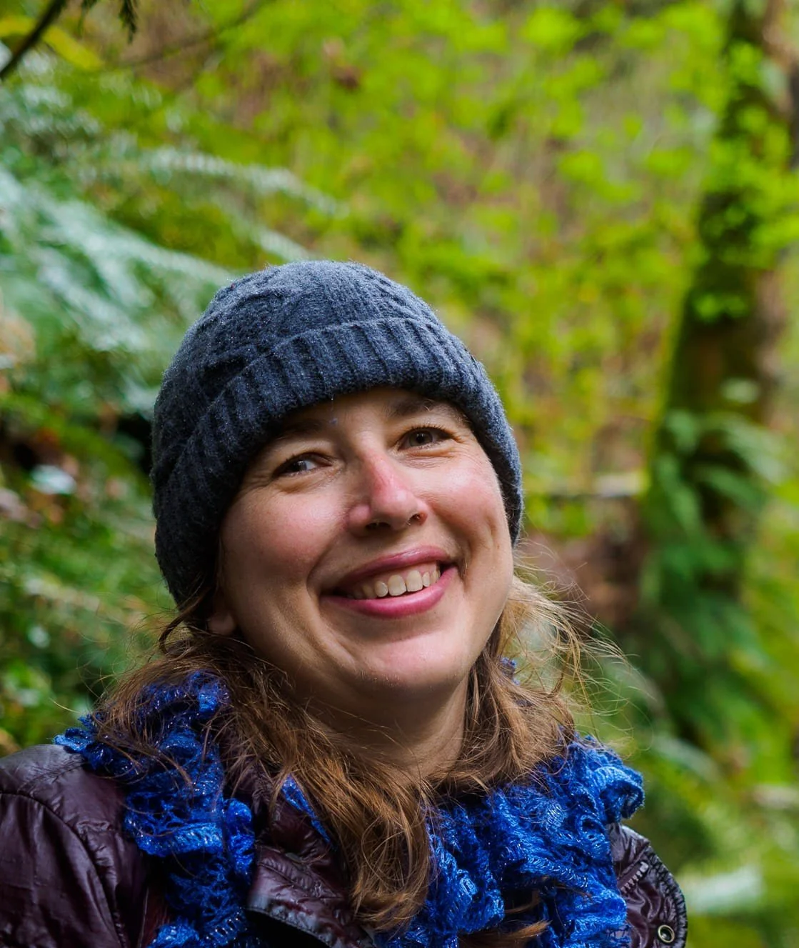 A woman smiling outdoors in a forest, wearing a blue knit beanie and a blue scarf.