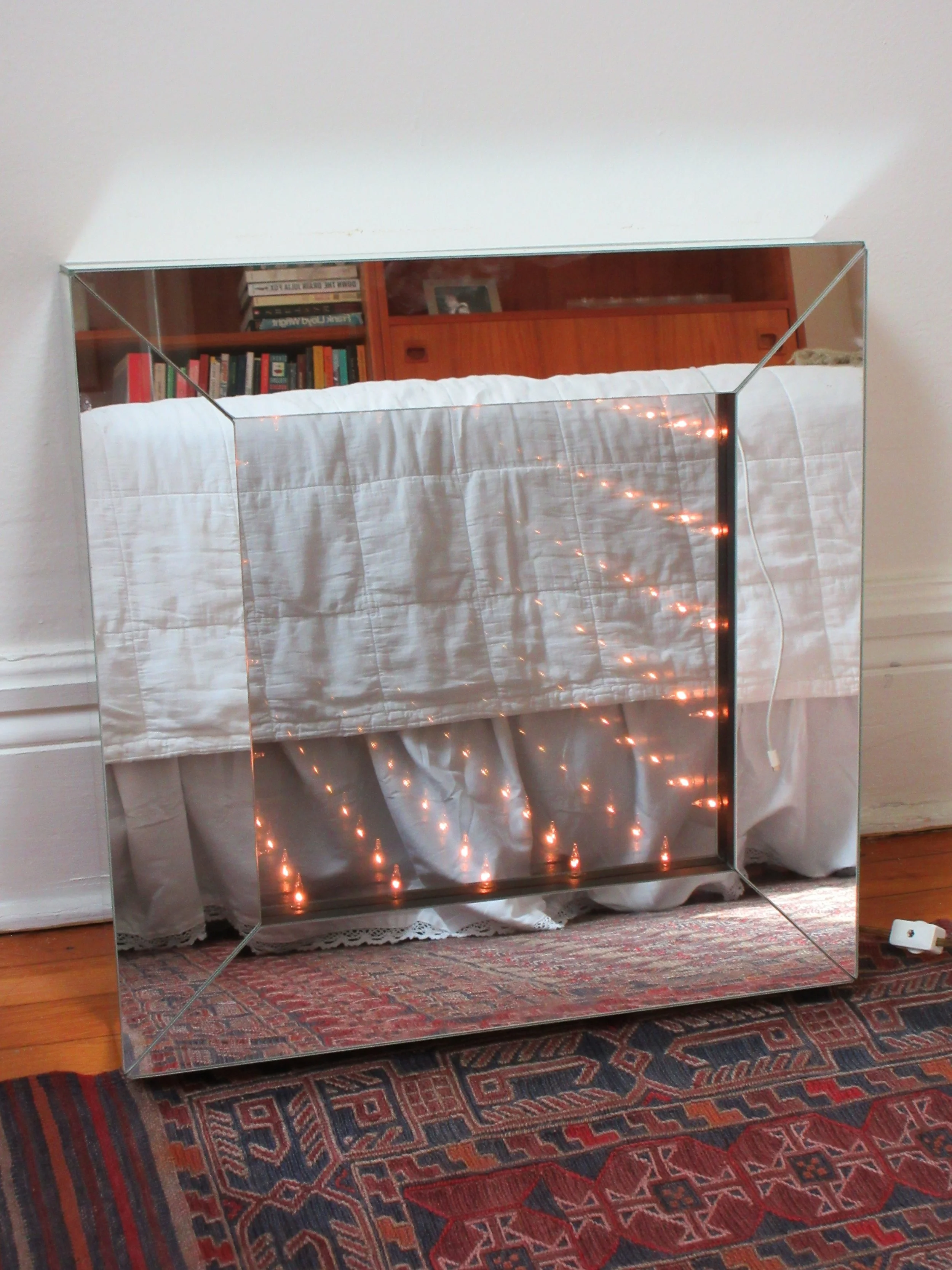 A large rectangular mirror with a reflective surface, placed on a wooden floor, reflects a bookcase and a bed with white bedding, a red wooden headboard, and a framed photo. The mirror has a silver frame and stands against a plain beige wall.