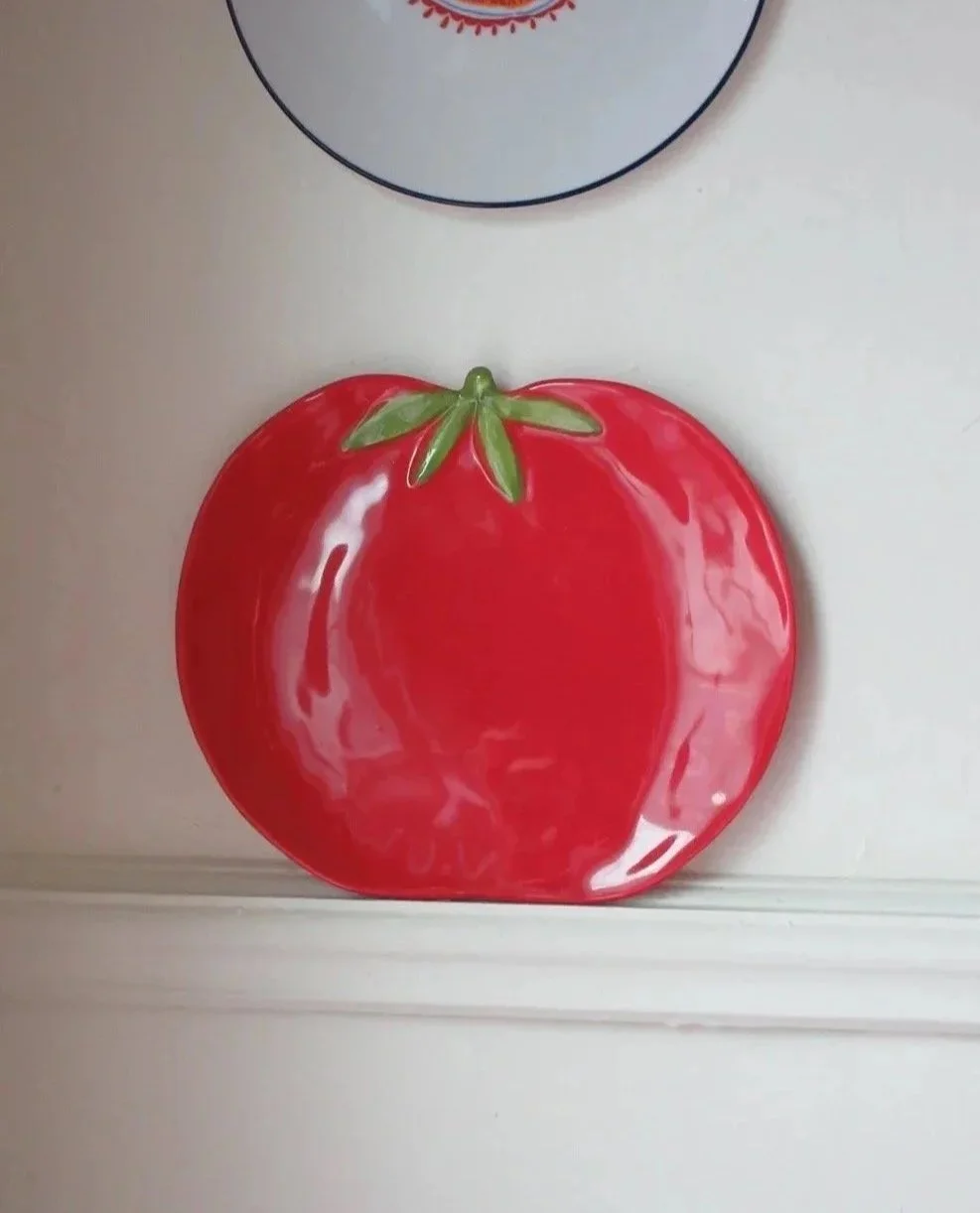 A ceramic plate shaped and painted like a red tomato, with green leaves at the top, sitting on a white surface.