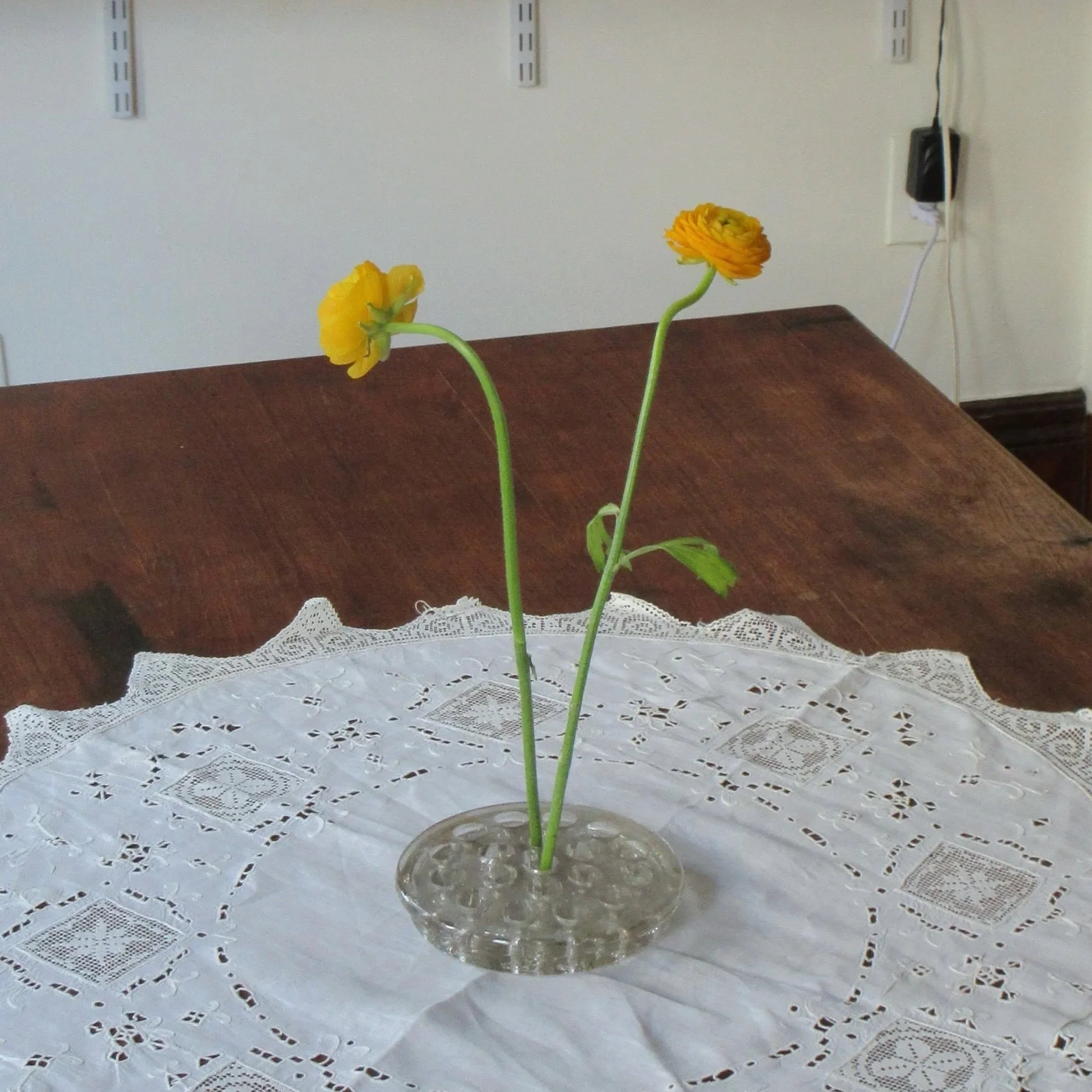 Two yellow flowers in a glass vase on a white lace tablecloth on a wooden table.