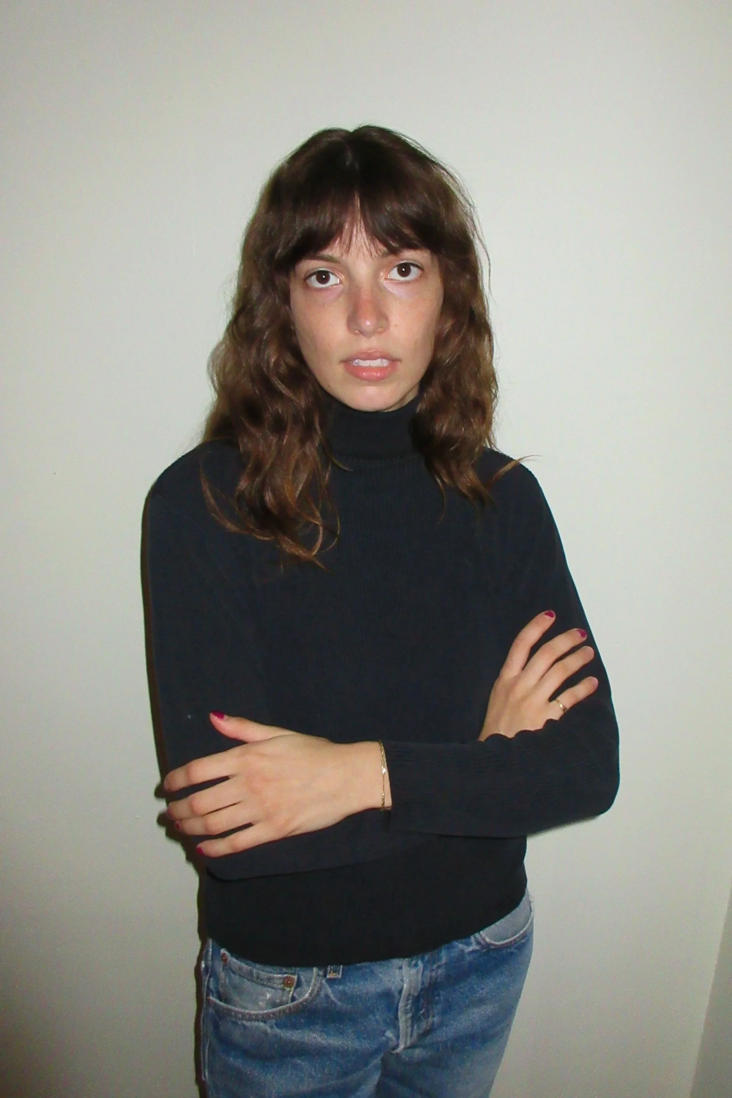 A woman with wavy brown hair, wearing a black turtleneck sweater and blue jeans, standing against a plain white wall with her arms crossed.