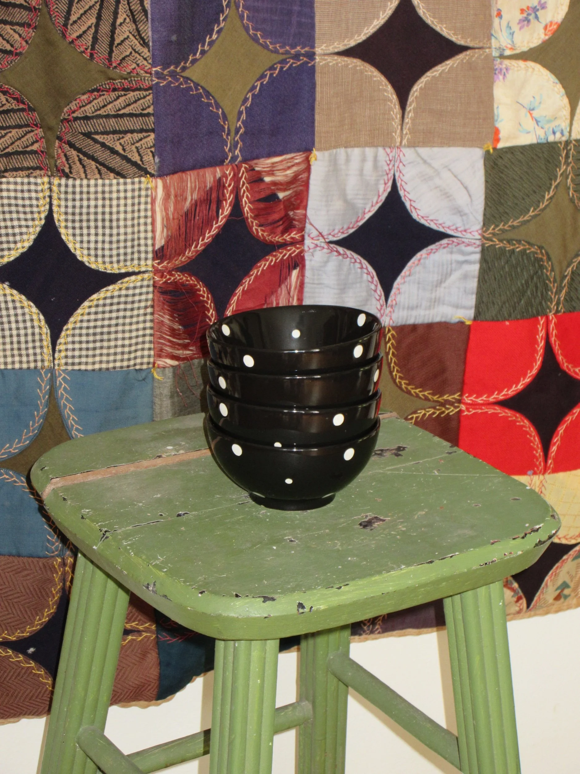 A small green wooden table with chipped paint, holding four black bowls with white polka dots, in front of a multicolored quilt backdrop.