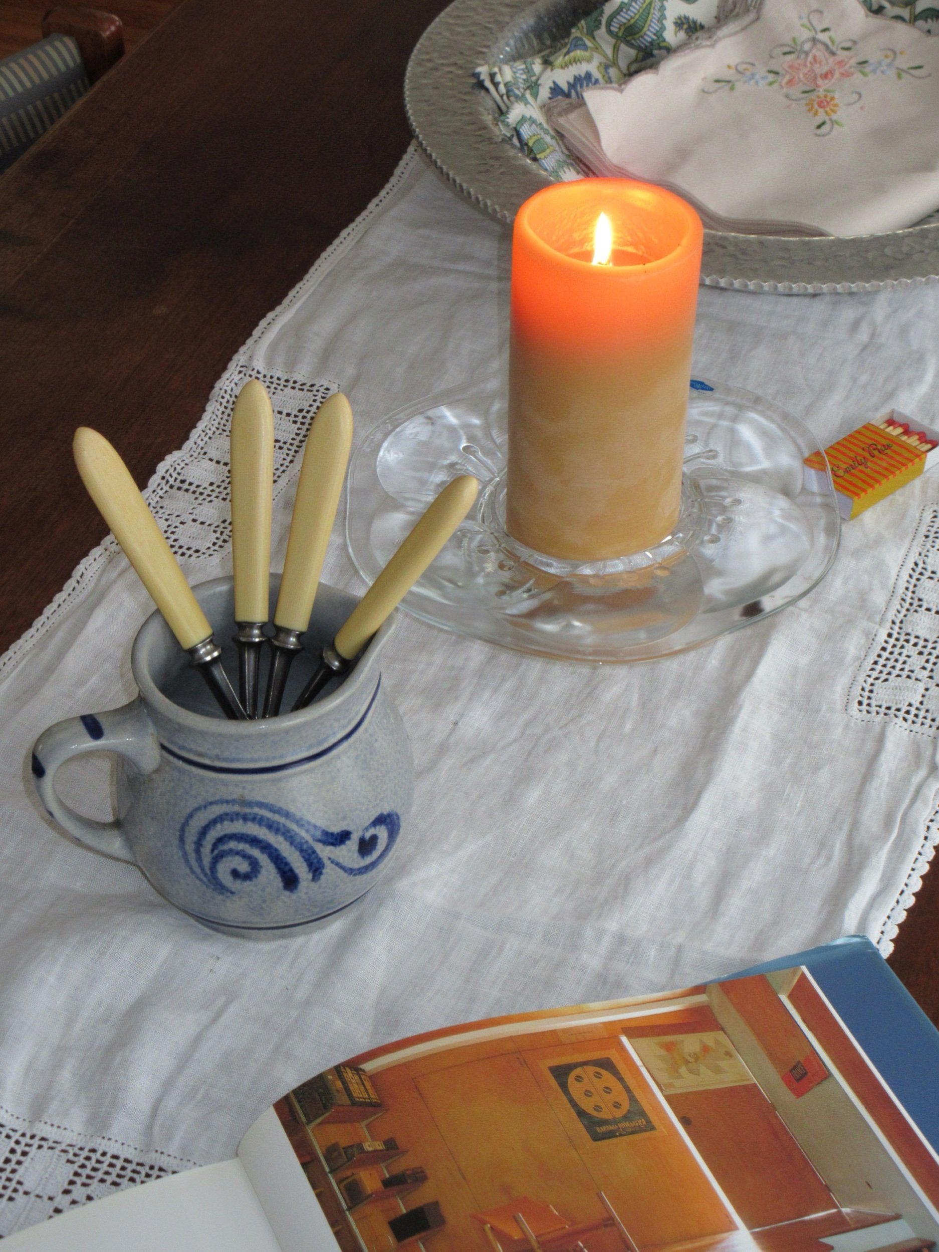 A table with a white cloth, a lit candle in a beige and white candlestick, a ceramic mug holding several knives with cream-colored handles, a partially open magazine, and a silver tray with cloth napkins.