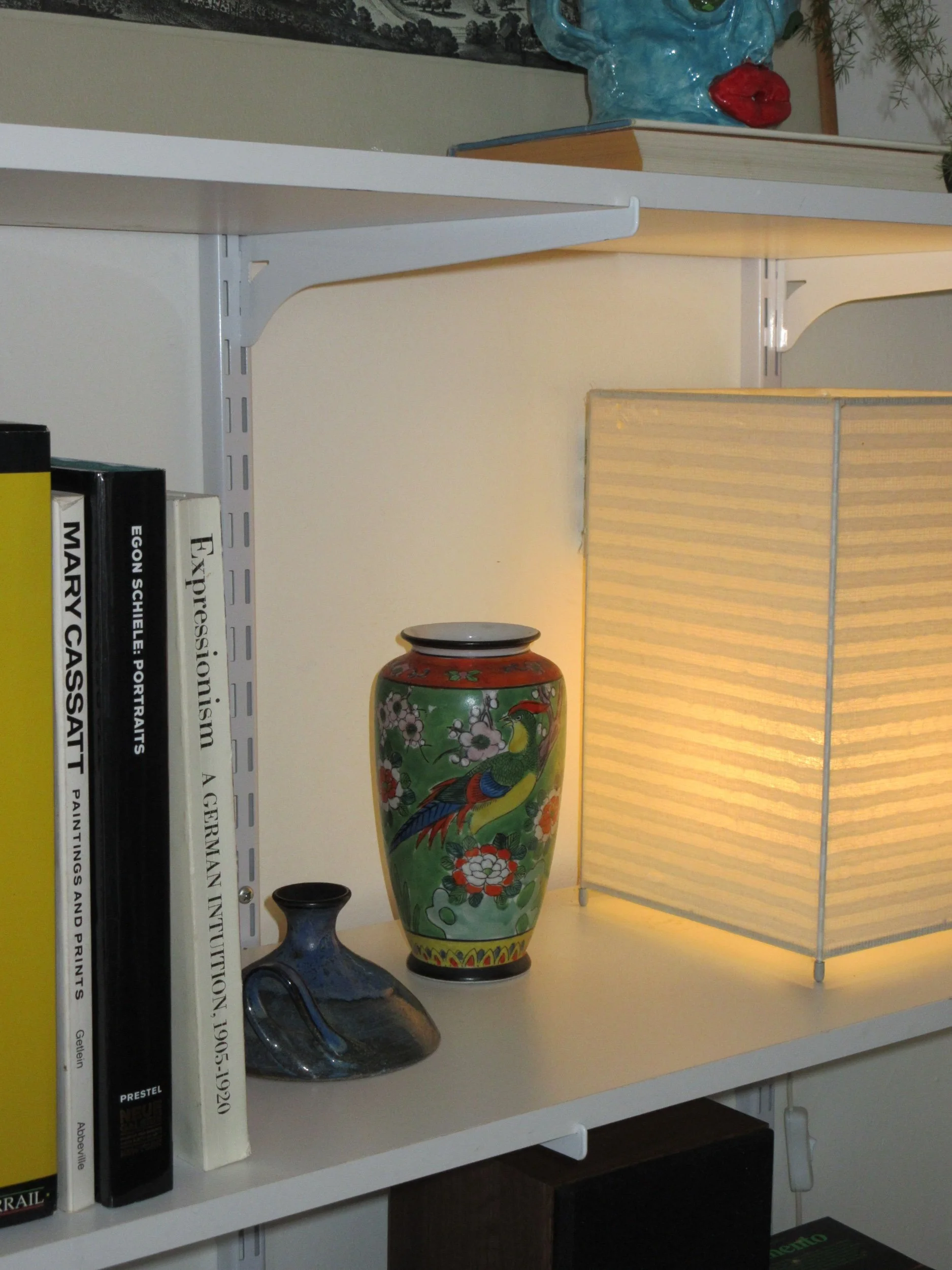 Decorative items on a white shelf including a colorful floral vase with a bird design, a small dark blue vase, a beige lampshade, books, and a blue decorative face sculpture with red lips in the background.