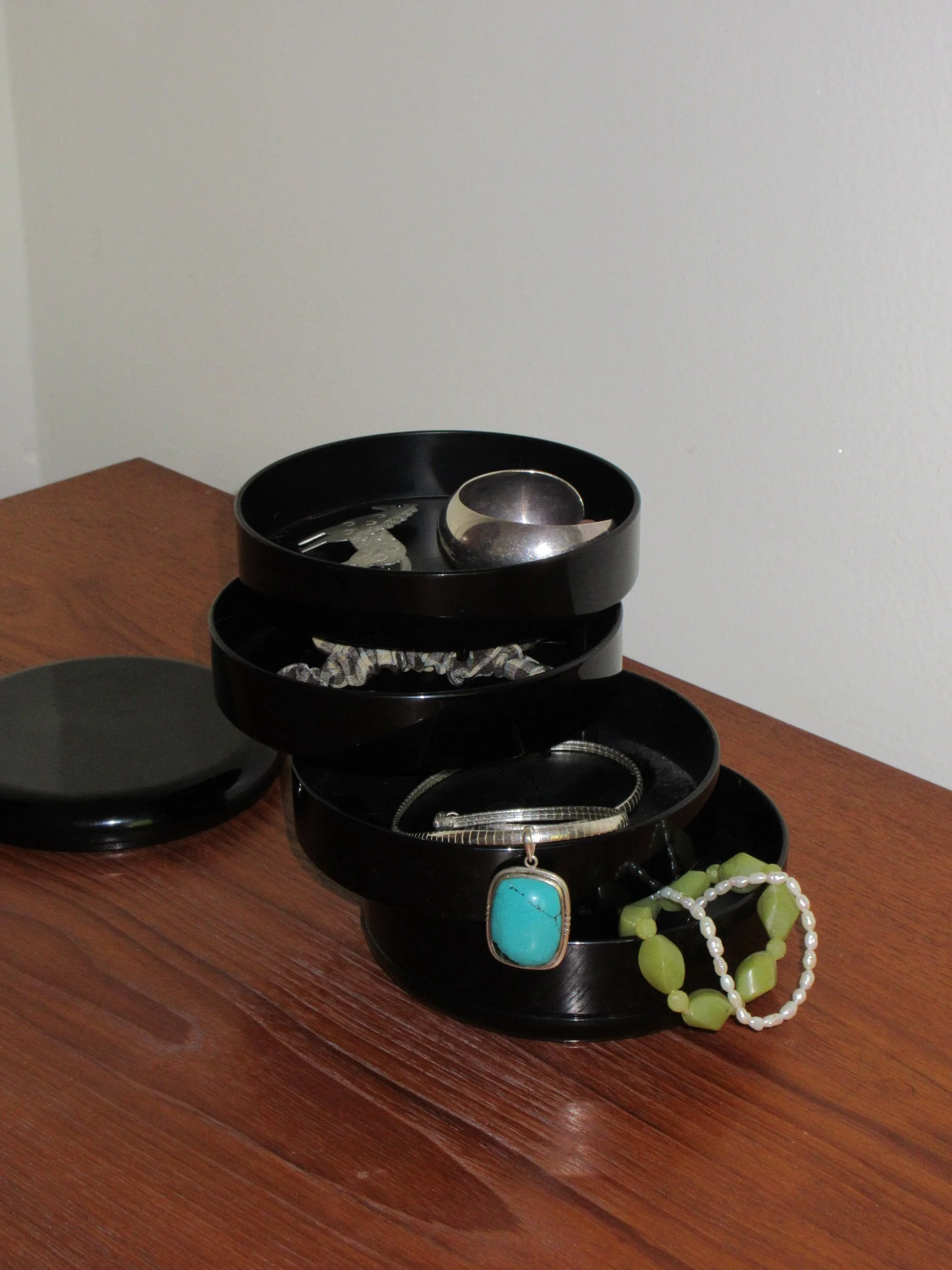 Stacked black jewelry containers with necklaces and bracelets, placed on a wooden surface against a plain wall.