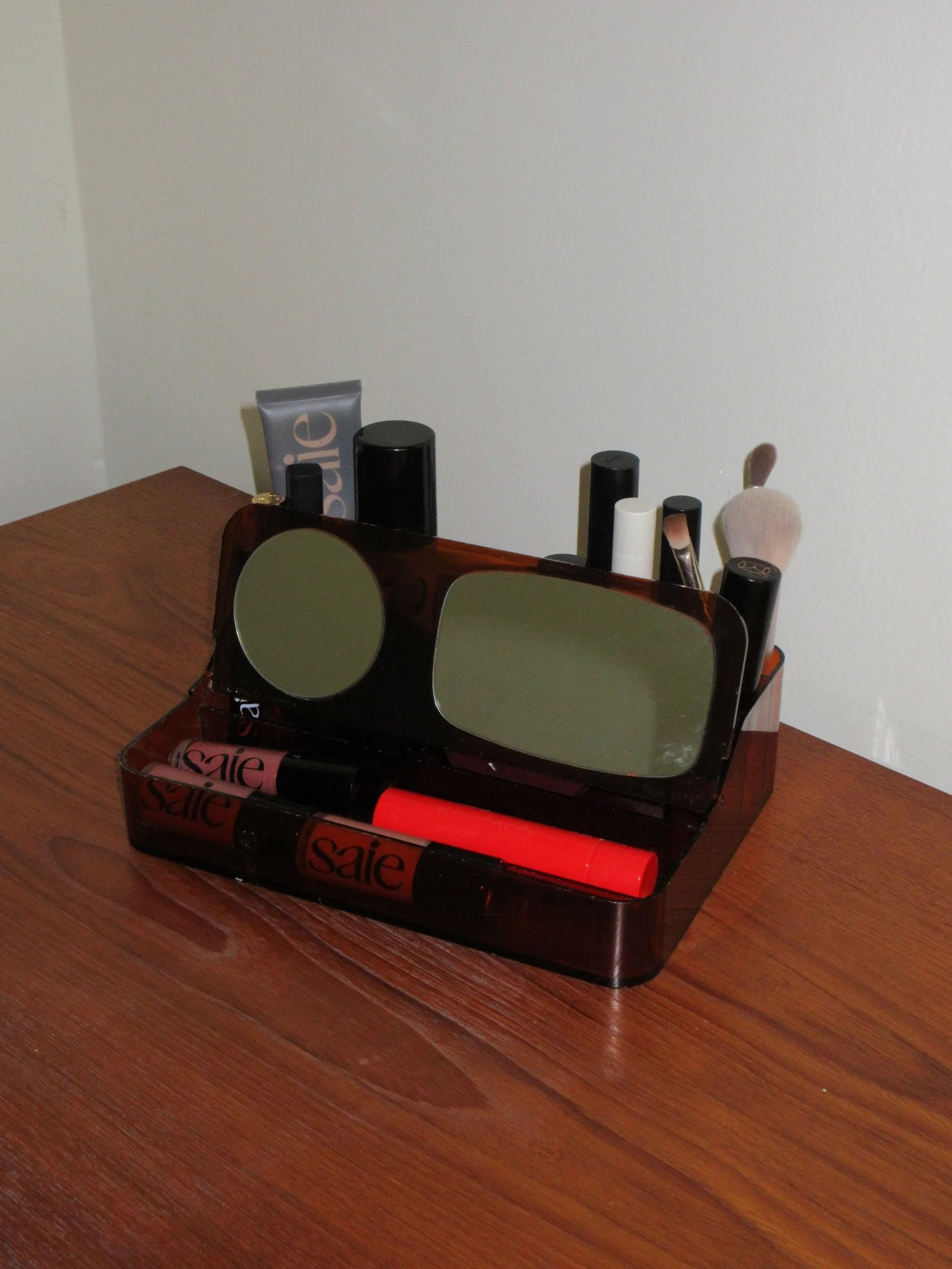 Makeup organizer with sunglasses, lip gloss, and brushes on a wooden surface.