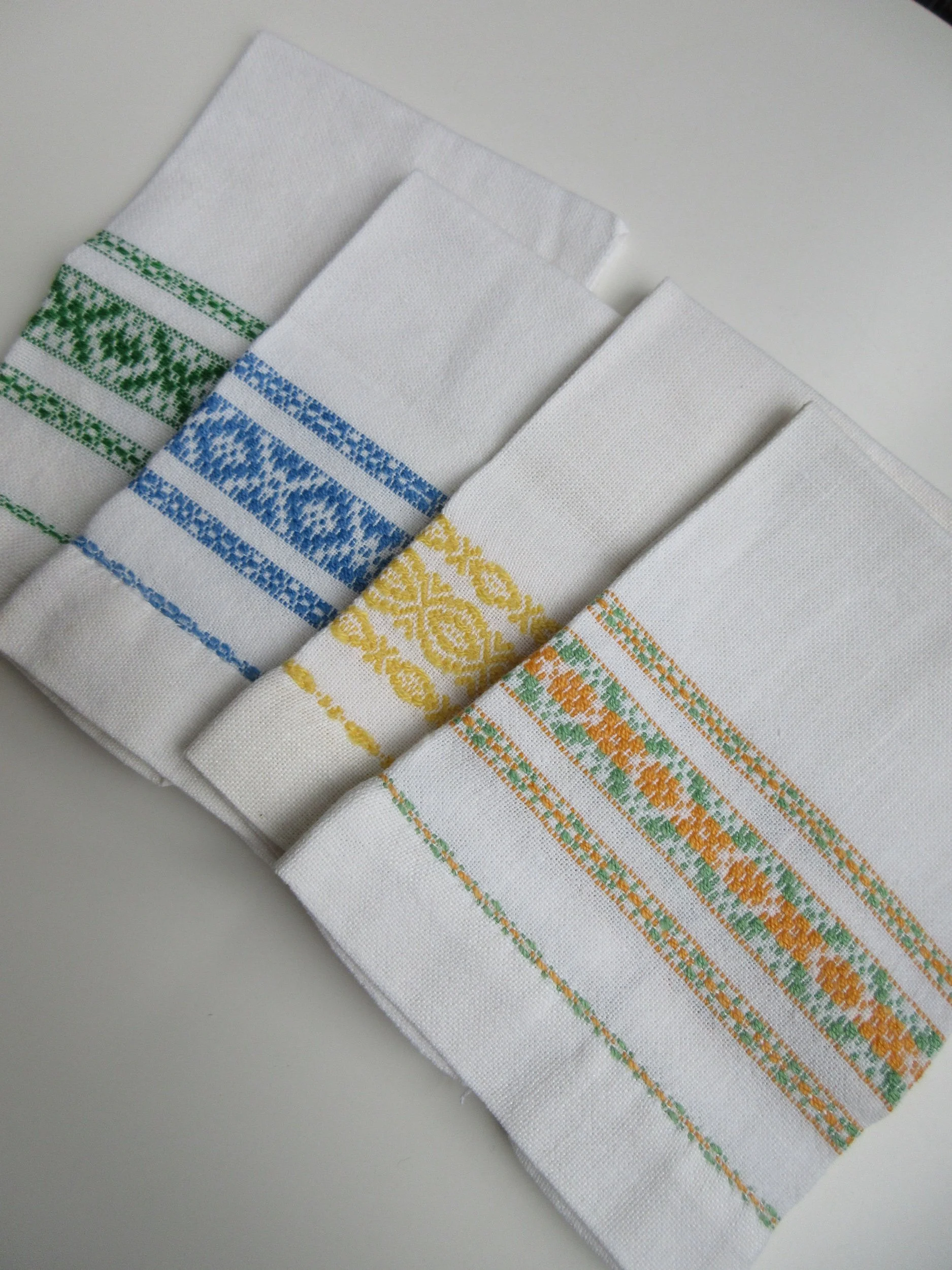 Four white kitchen towels with colorful embroidered patterns in green, blue, yellow, and orange, arranged side by side on a white surface.