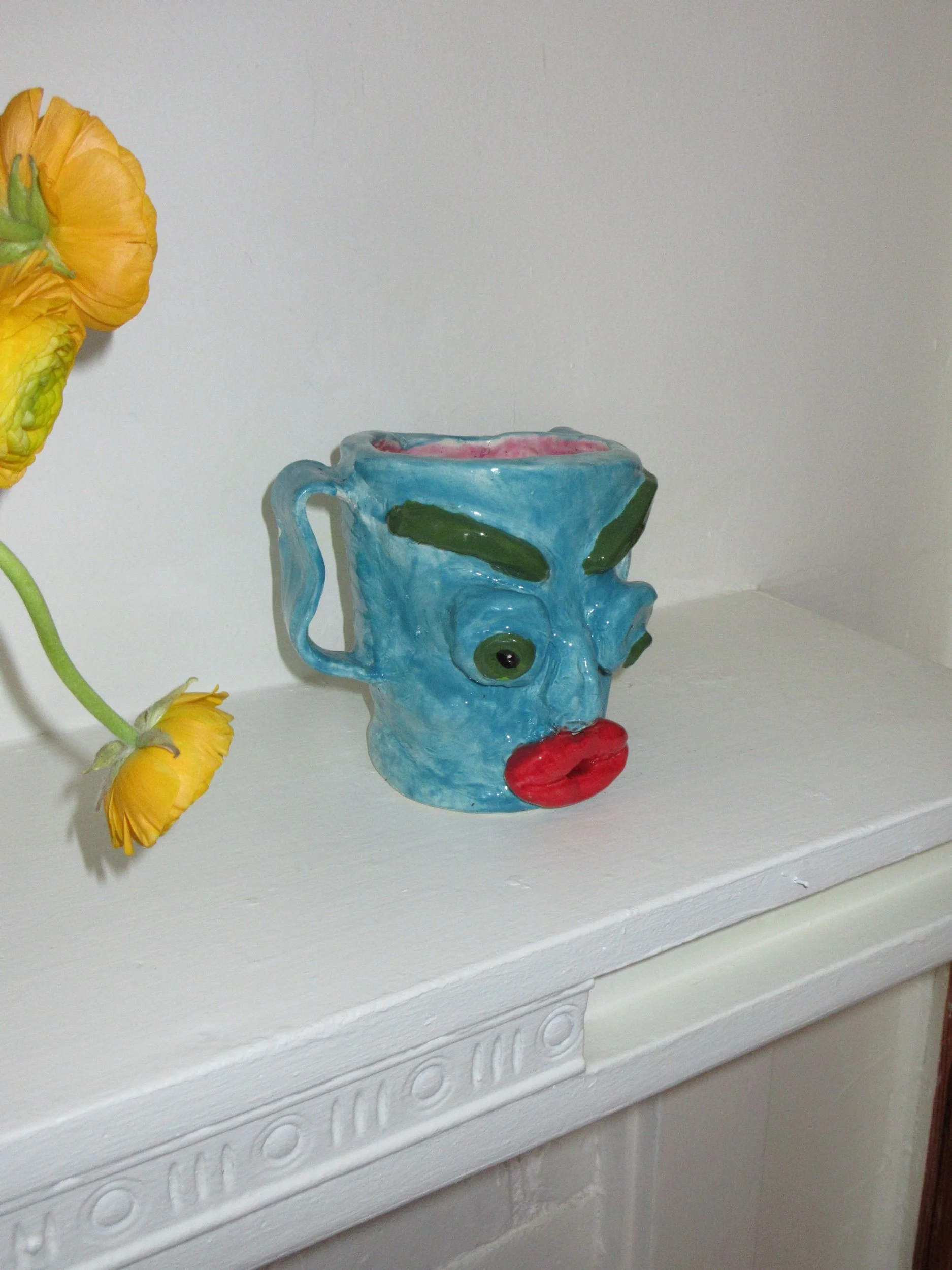 A ceramic mug shaped like a face with a blue glaze, red lips, green eyebrows, and green eyes, placed on a white surface next to yellow flowers.