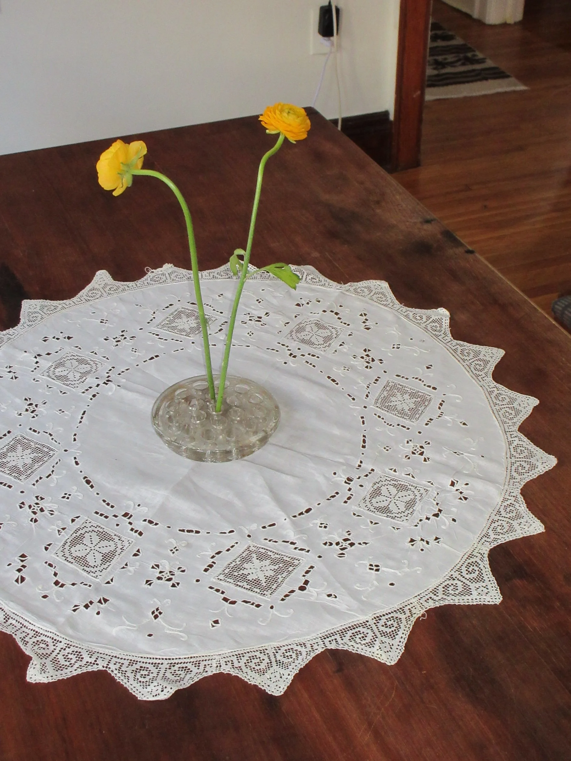 Yellow flowers in a clear glass vase on a white lace tablecloth on a wooden table.