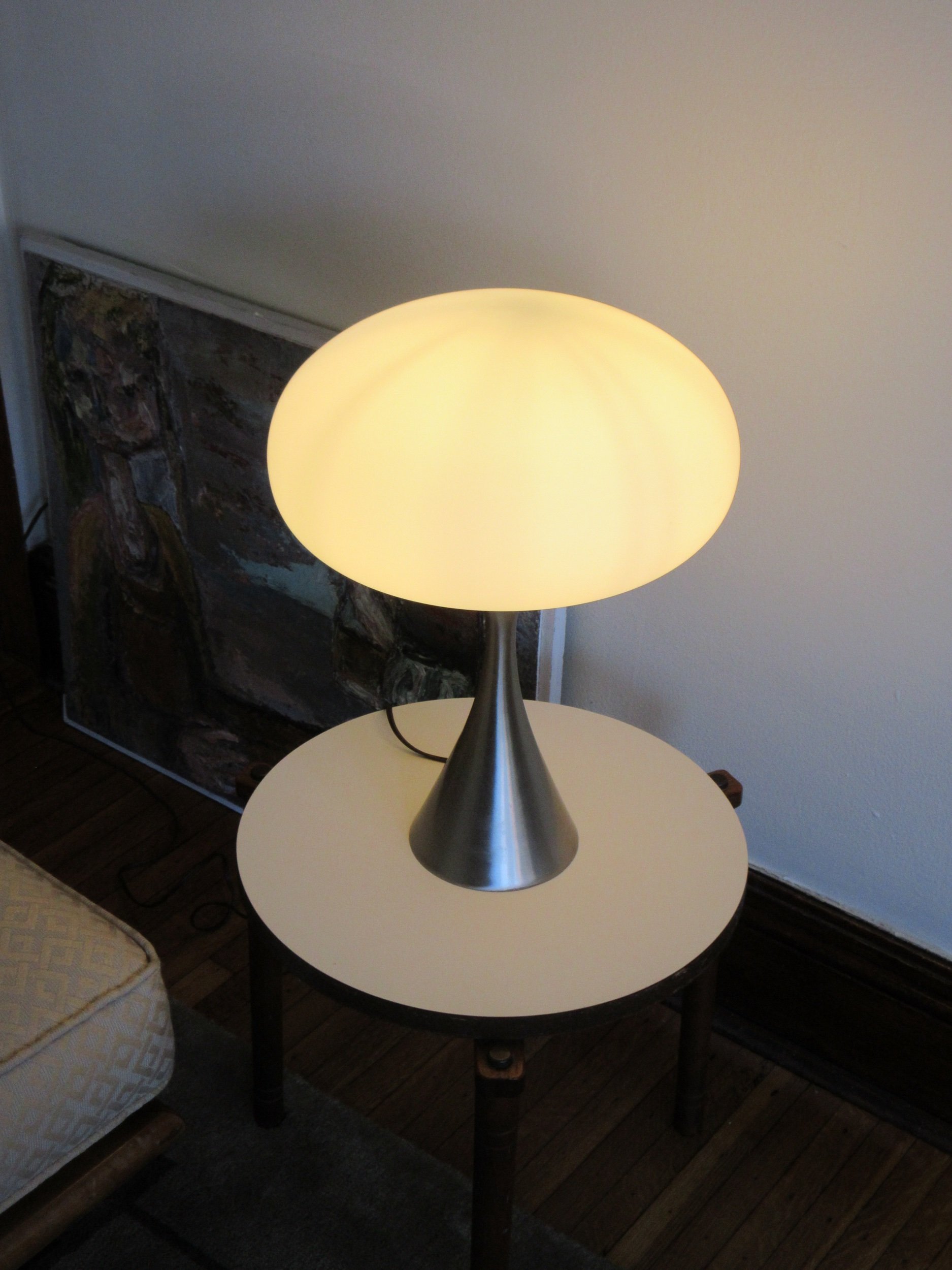A modern table lamp with a brushed metal cone-shaped base and a large, round, yellowish lampshade, placed on a small white round table in a room with a painting and wooden floor.