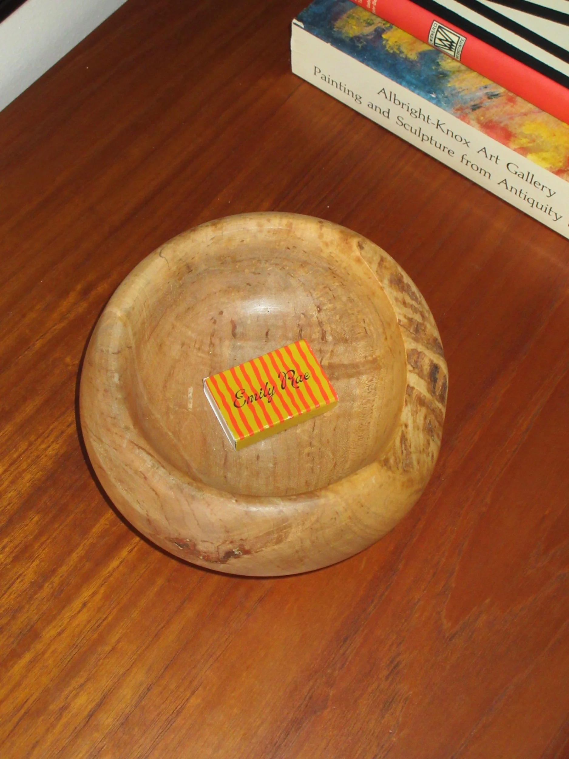 A small rectangular box with a yellow and red striped pattern labeled 'Emily Rae' inside a wooden bowl on a wooden surface, with books in the background.