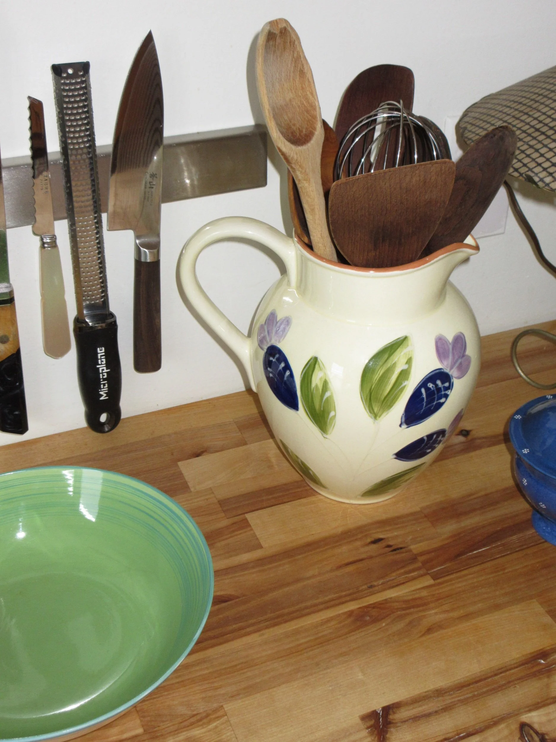 Kitchen utensils including knives and graters hanging on a strip above a wooden countertop, with a ceramic pitcher holding wooden spoons, a whisk, and a wooden spatula.