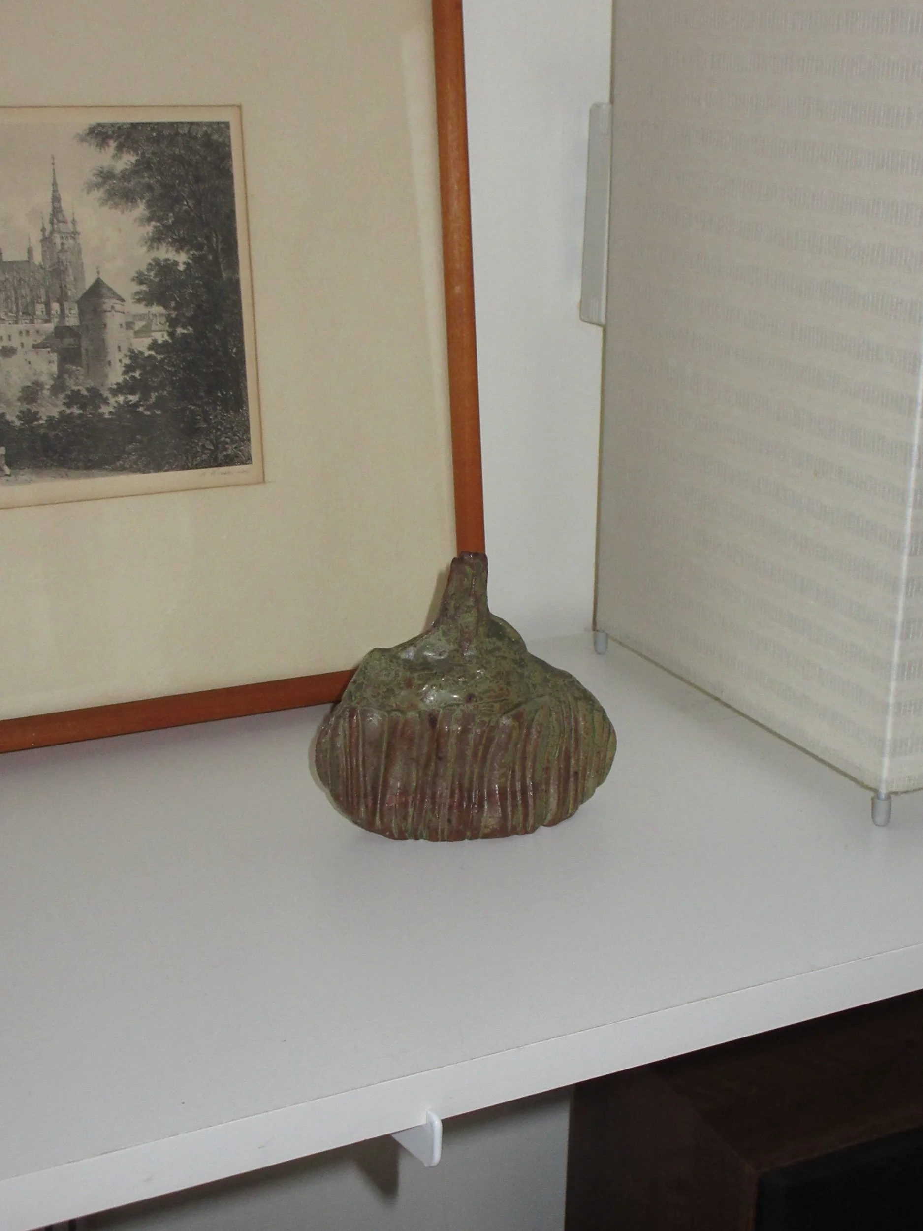 Ceramic sculpture shaped like a pumpkin placed on a white shelf, with framed artwork featuring a castle or historical building and a tree behind it, and a white room divider on the right