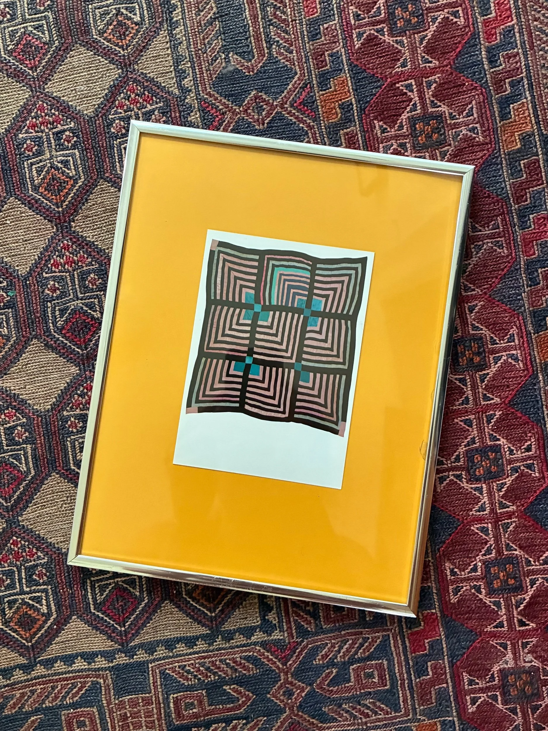 Framed geometric artwork resting on a vibrant, patterned rug with red, blue, and beige colors.
