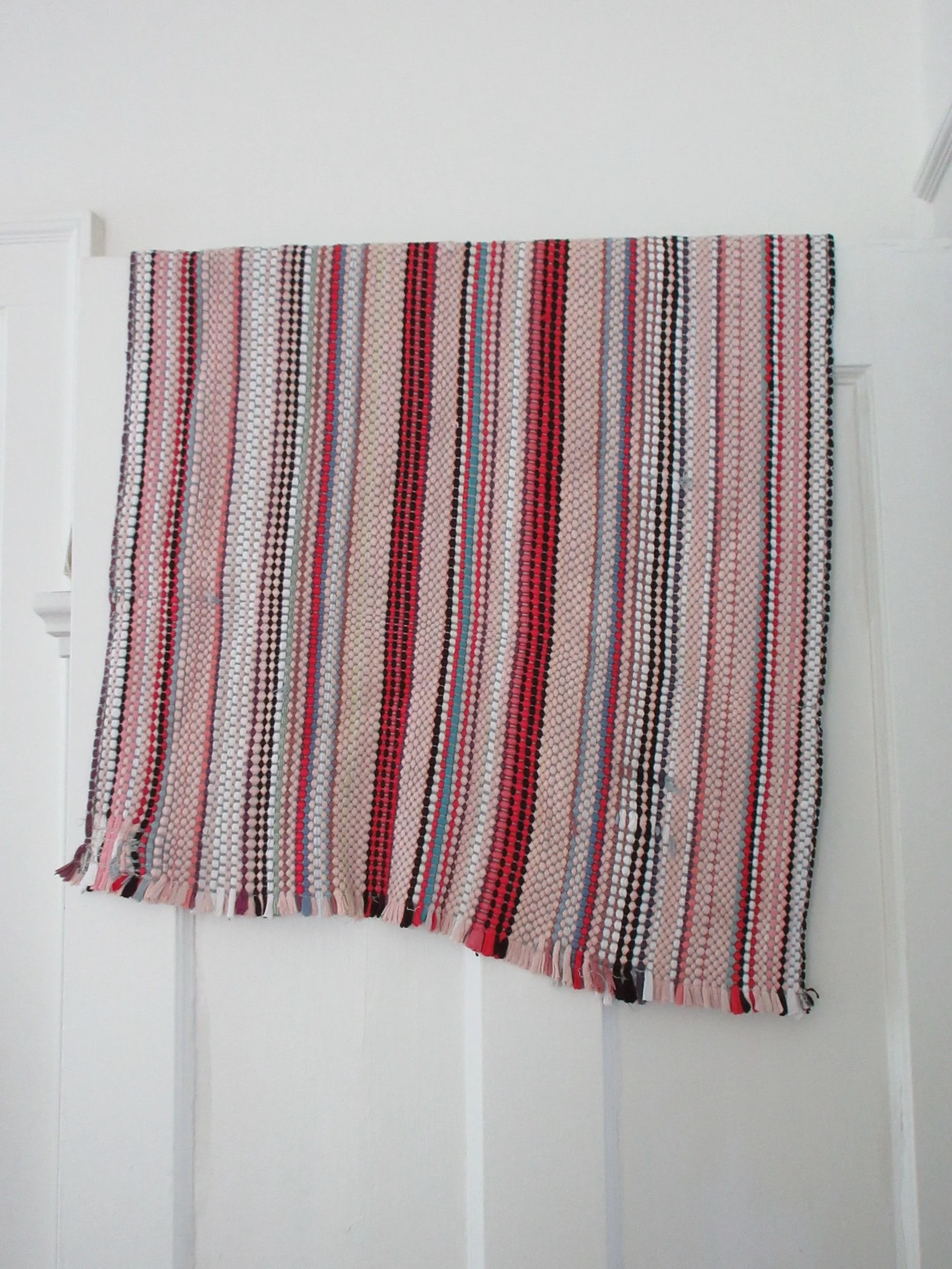 Colorful woven rug with red, pink, black, white, and teal striped pattern hanging on a white wall.