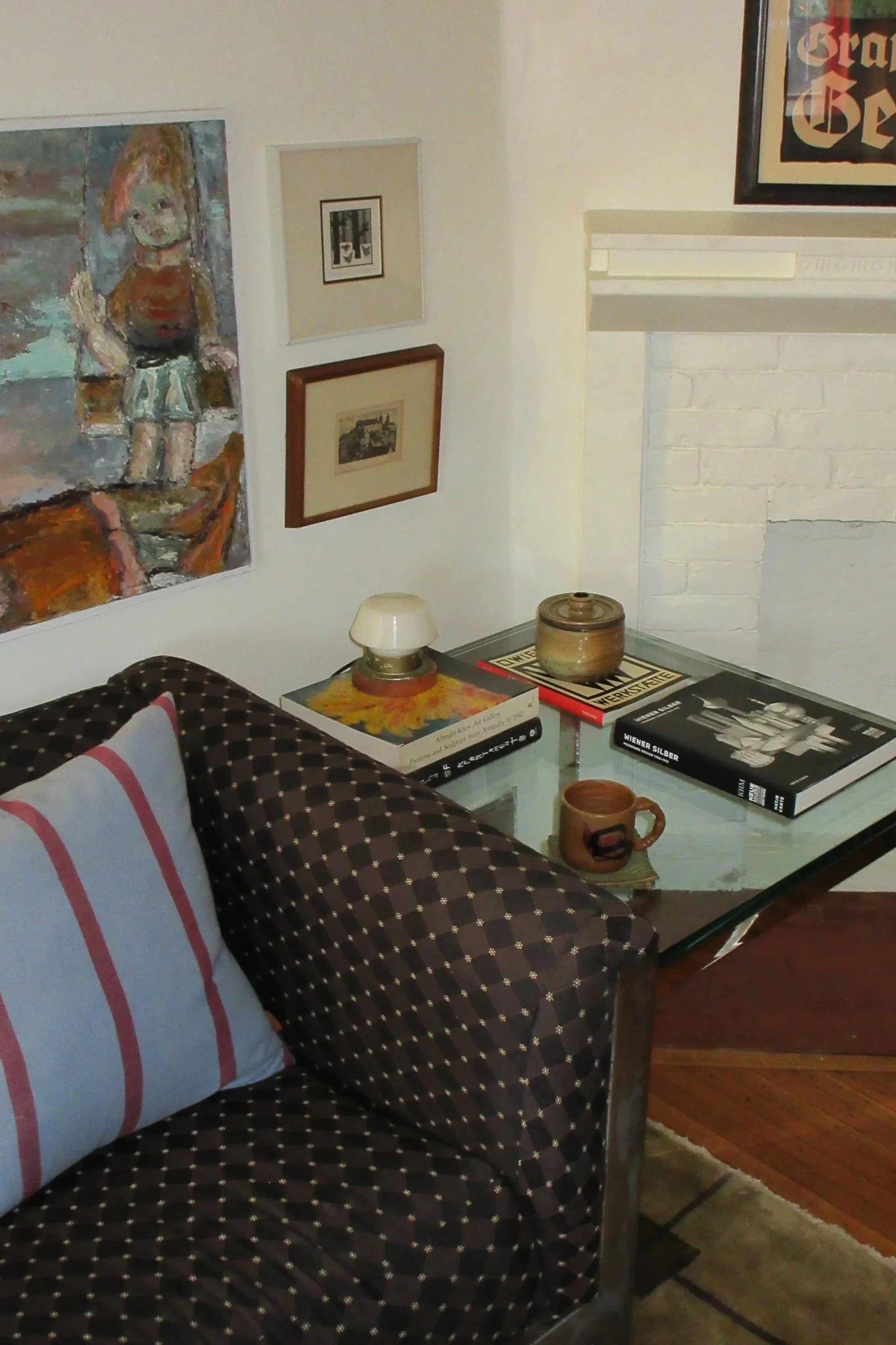 Living room corner with a dark patterned sofa, a glass-top table with books and ceramics, a wall with framed artwork and a textured white brick fireplace.